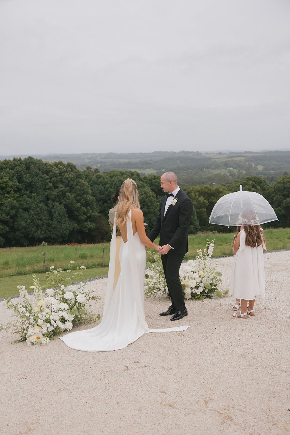 Bride and groom exchange vows on hilltop with flower girl holding clear umbrella nearby.