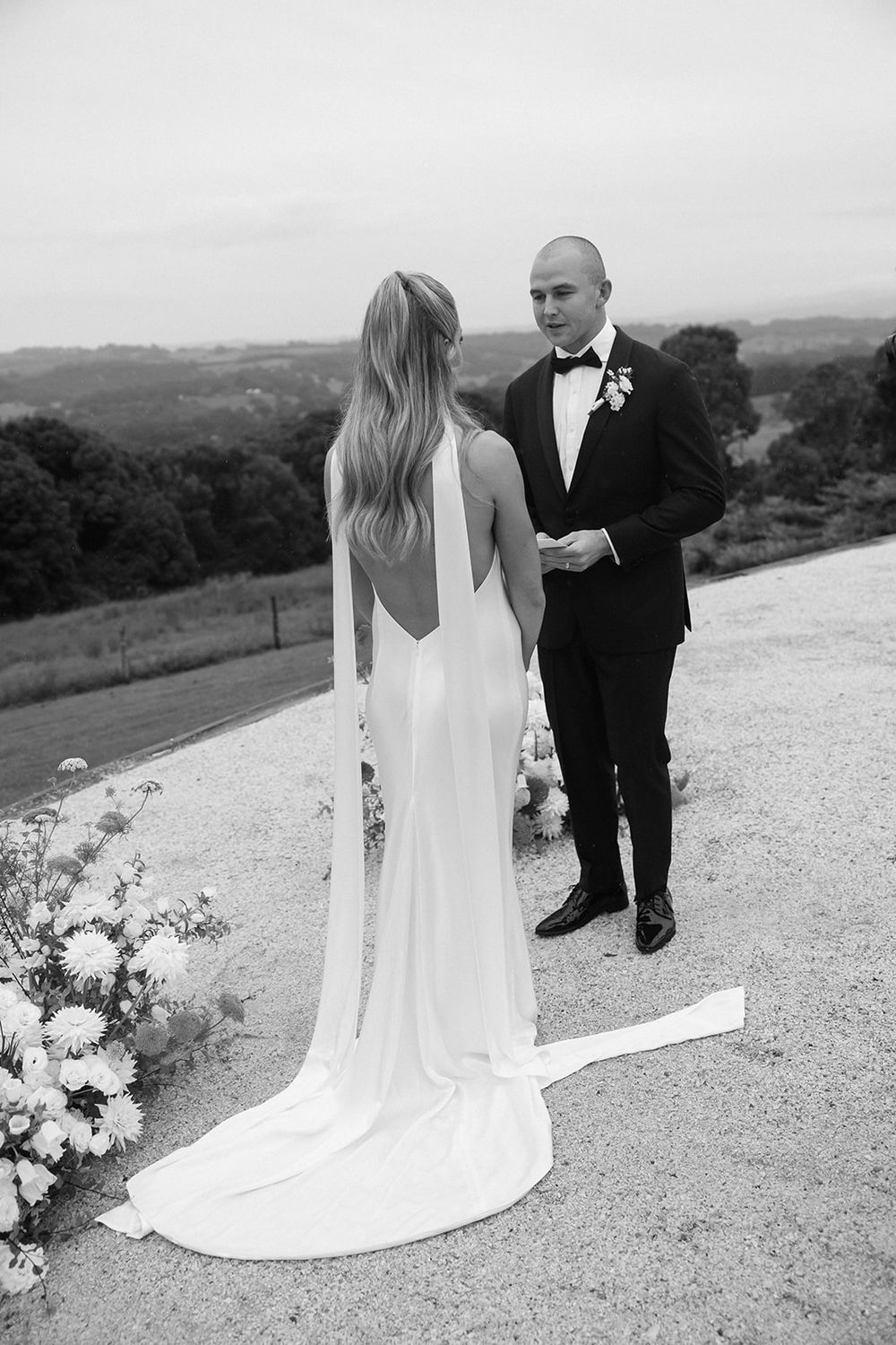 Elegant bride in backless gown stands with groom against scenic hillside backdrop.