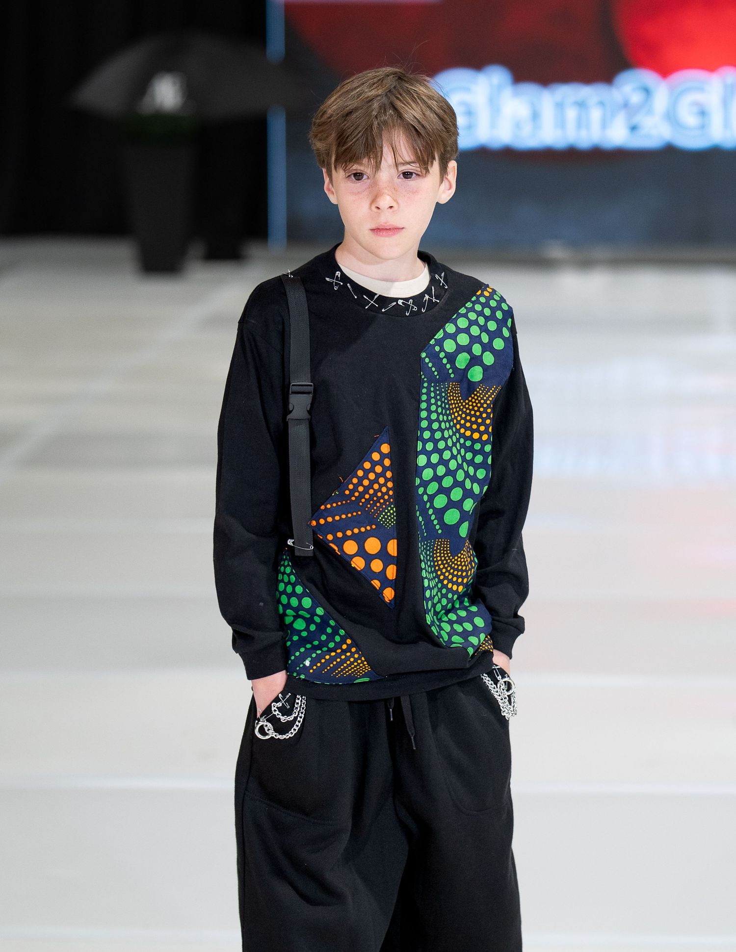 Model walks runway in black sweatshirt with geometric patterns in green and orange worn with dark baggy pants.