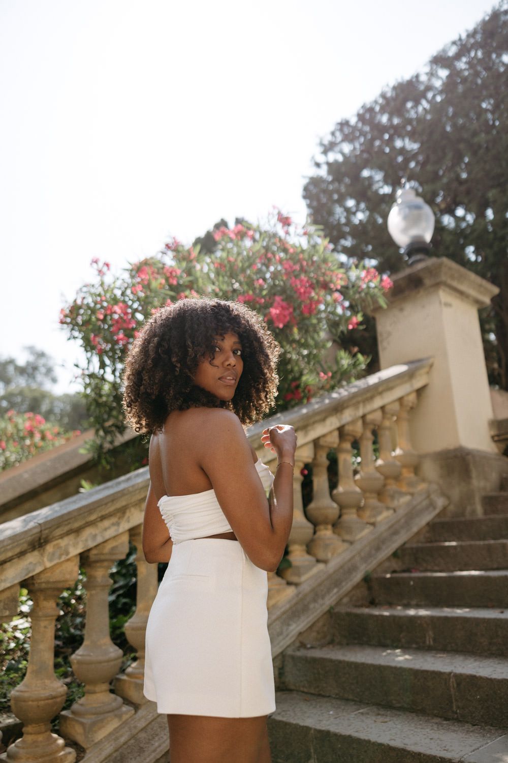 A person in a white dress poses on ornate stone steps with blooming red flowers in the background.