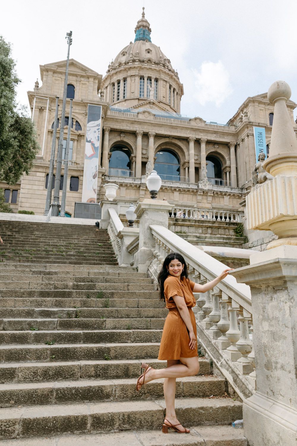 A person in brown dress poses on the grand staircase of the National Palace of Montjuic in Barcelona.