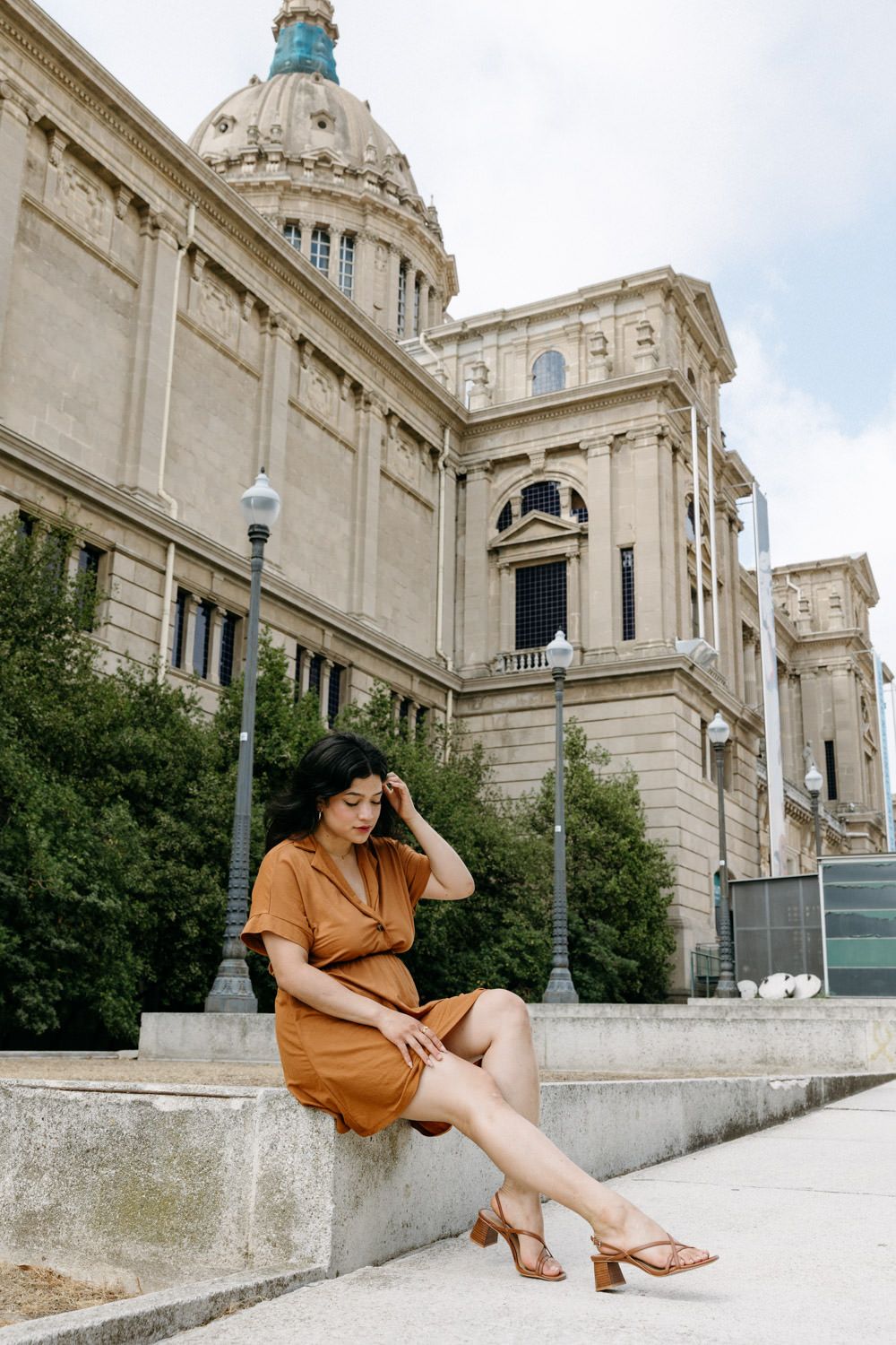 A person in rust-colored dress sits elegantly on stone steps near a domed classical building.