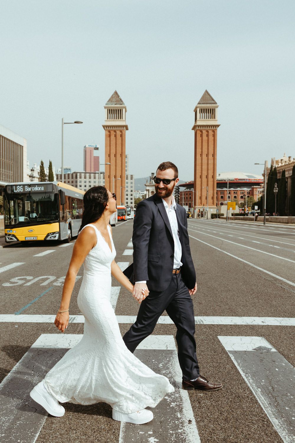 A couple in wedding attire walks together on a street with iconic towers in Barcelona.