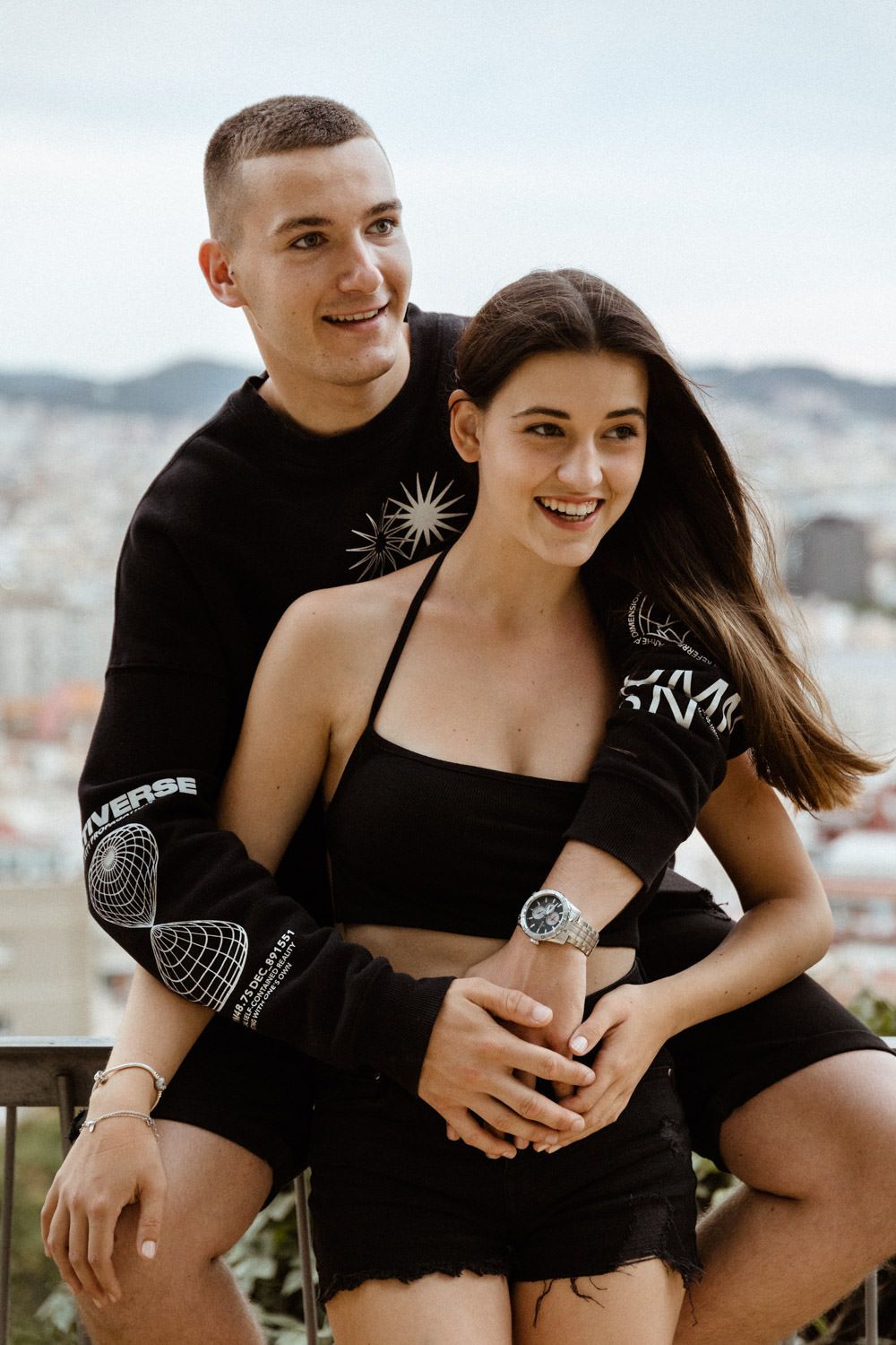 Young couple in matching black outfits embracing on balcony with city view.