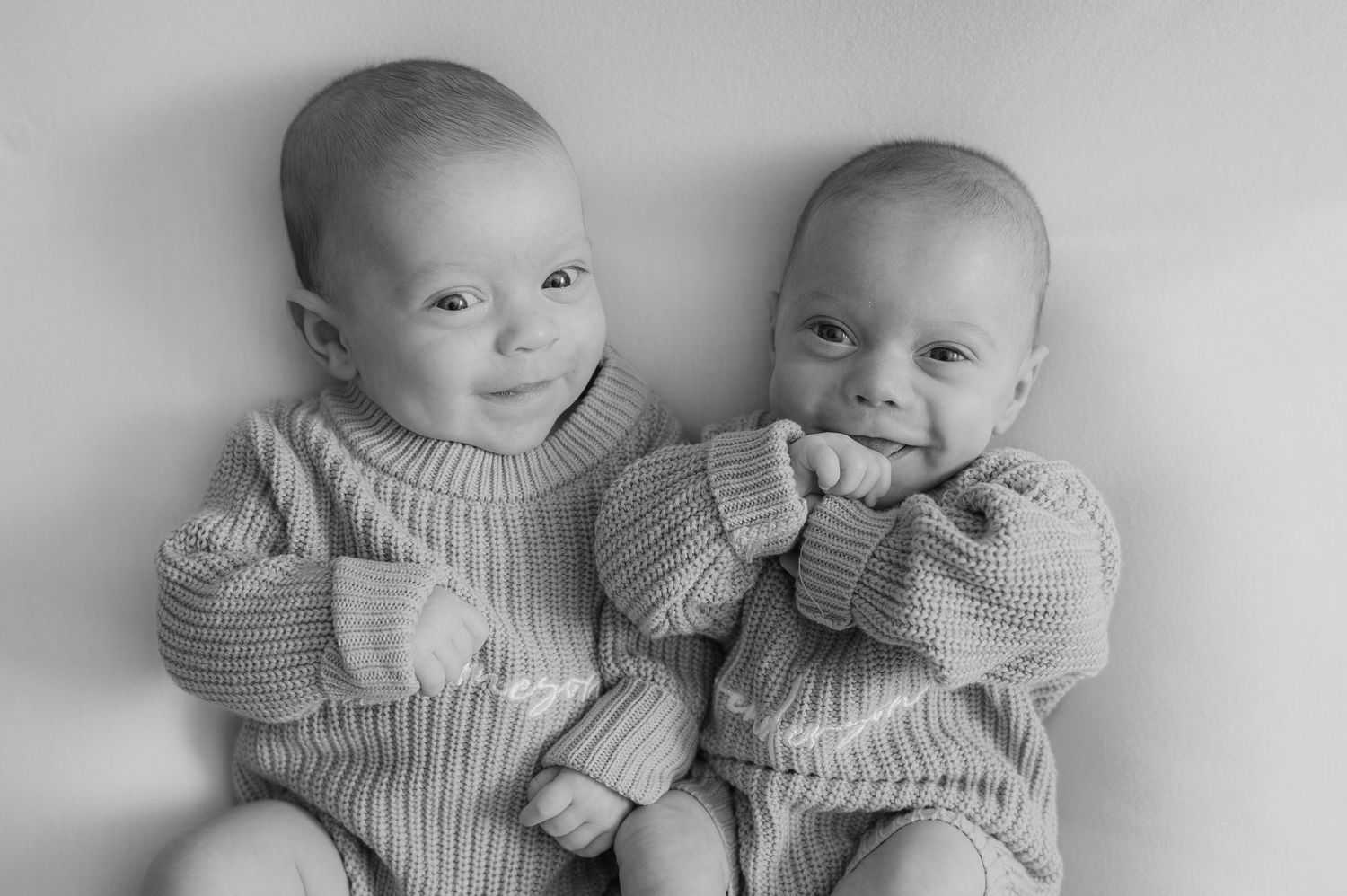 Black and white photo of two smiling babies in matching knit onesies lying side by side, likely twins.