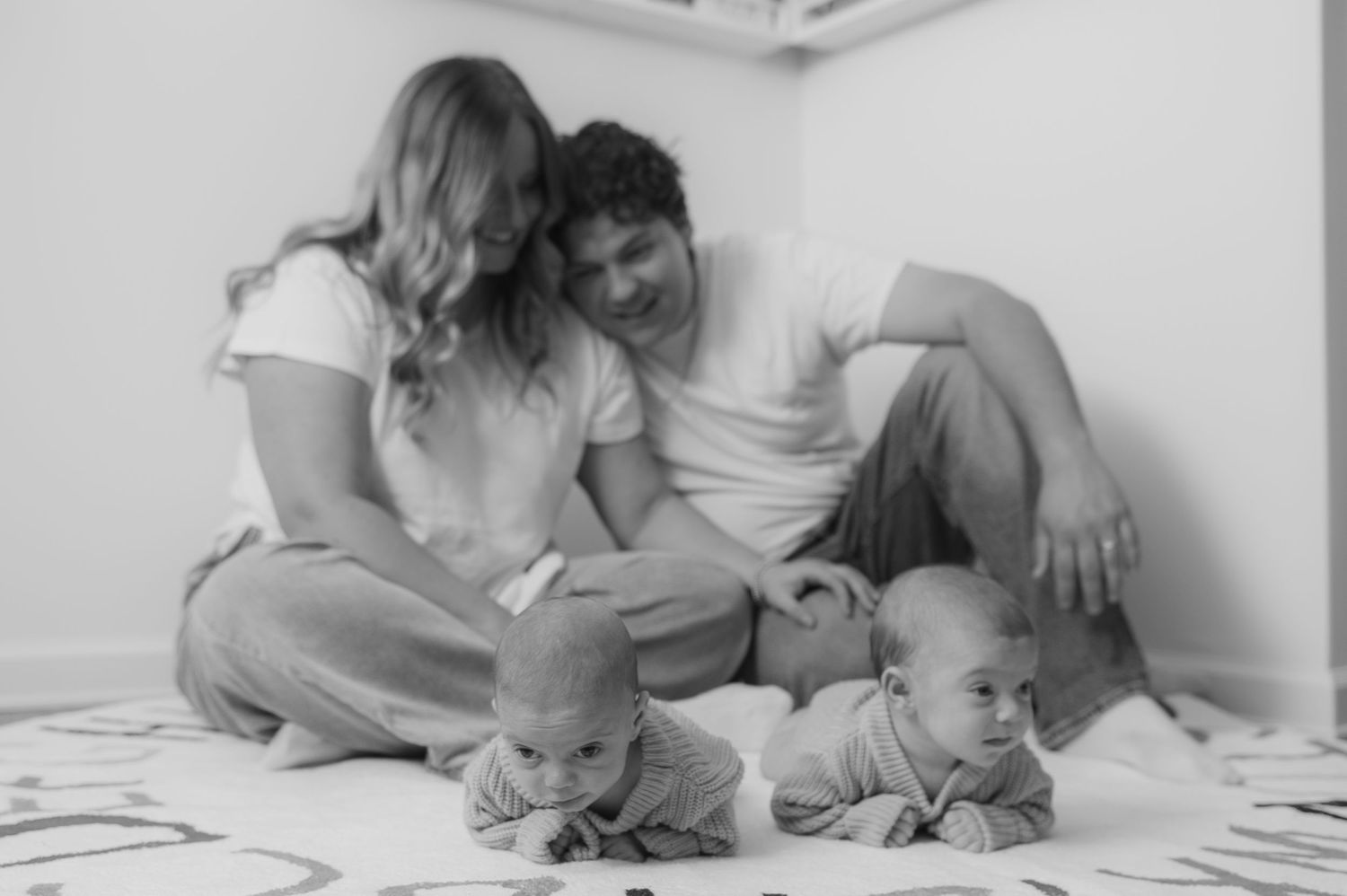 Black and white photo of parents smiling at their twin babies doing tummy time on a play mat.