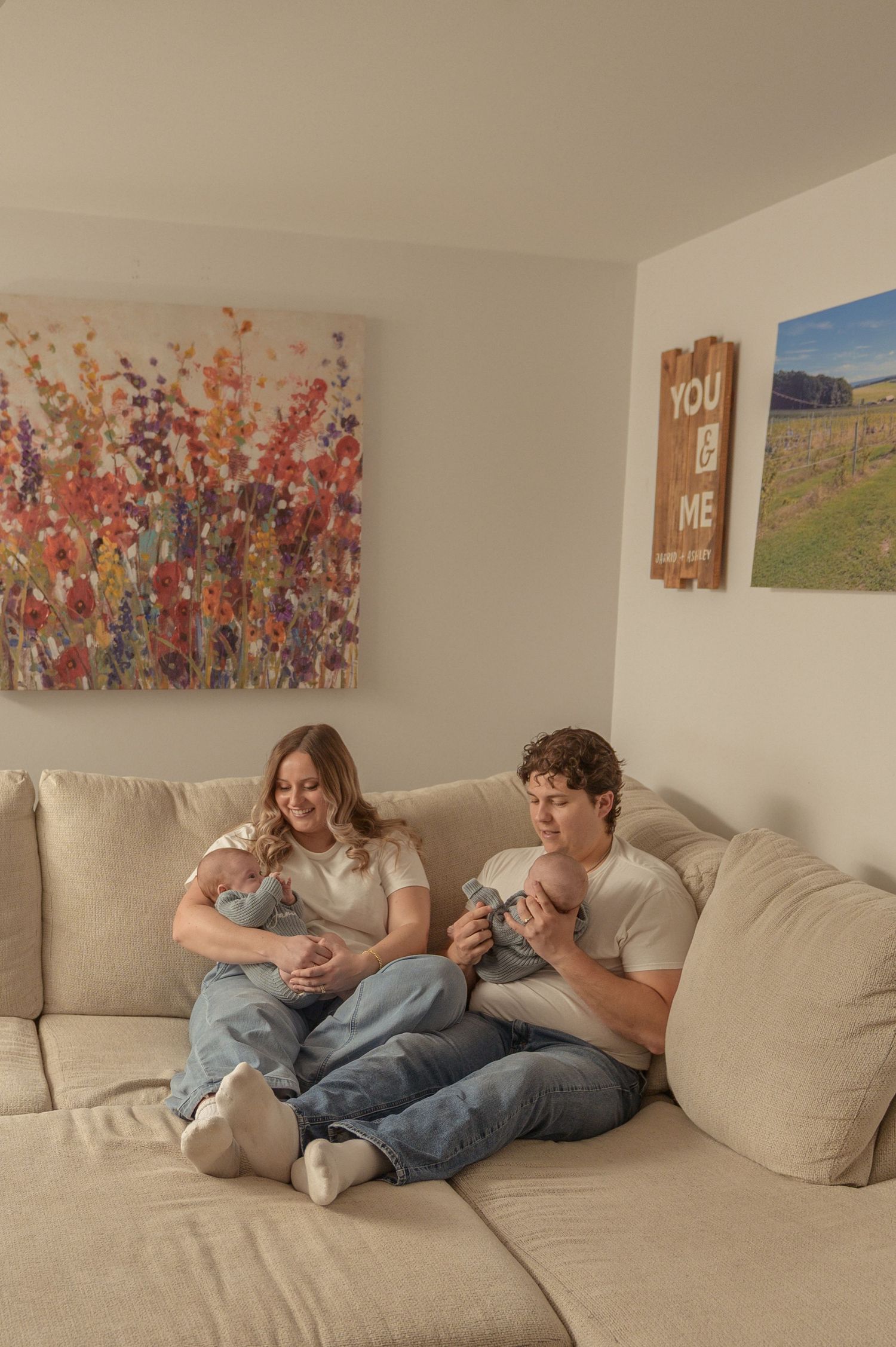 A young couple sits on a beige sectional sofa, lovingly holding their newborn baby in a cozy living room.