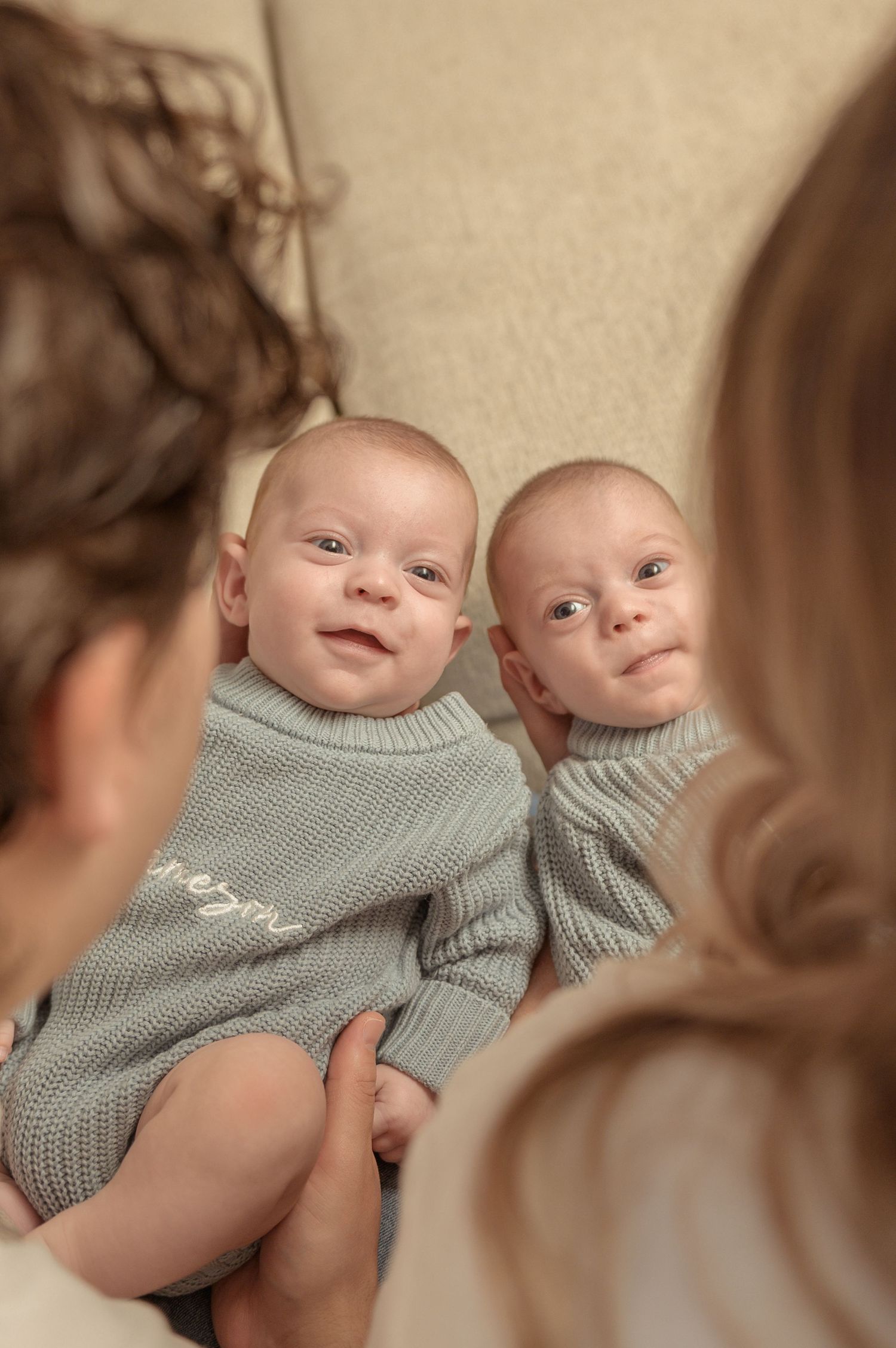 Two smiling twin babies in matching sage green knit outfits being held by a person, viewed from behind.