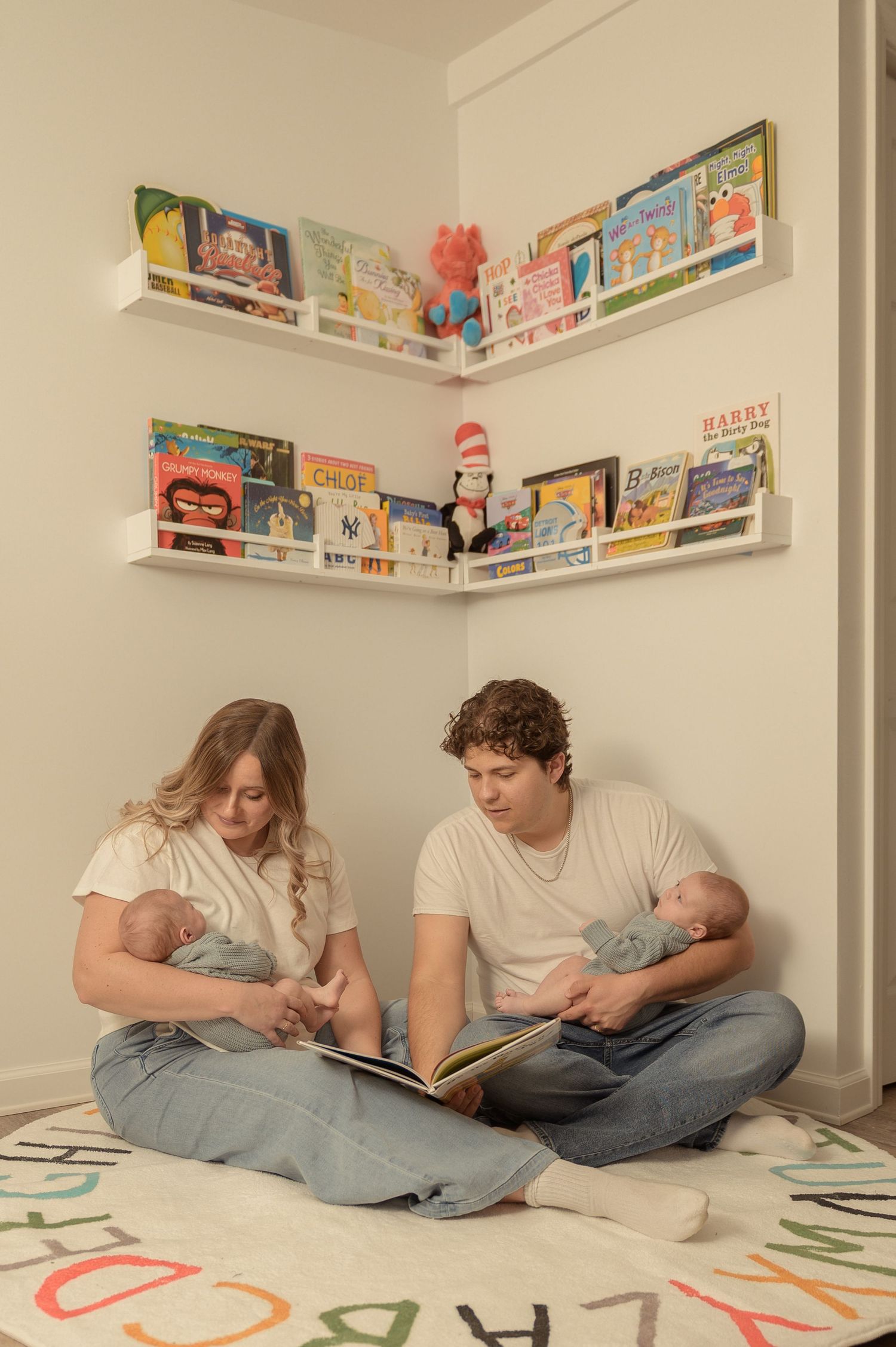 Parents sit on alphabet rug reading to newborn twins in nursery corner with colorful children's book shelves.