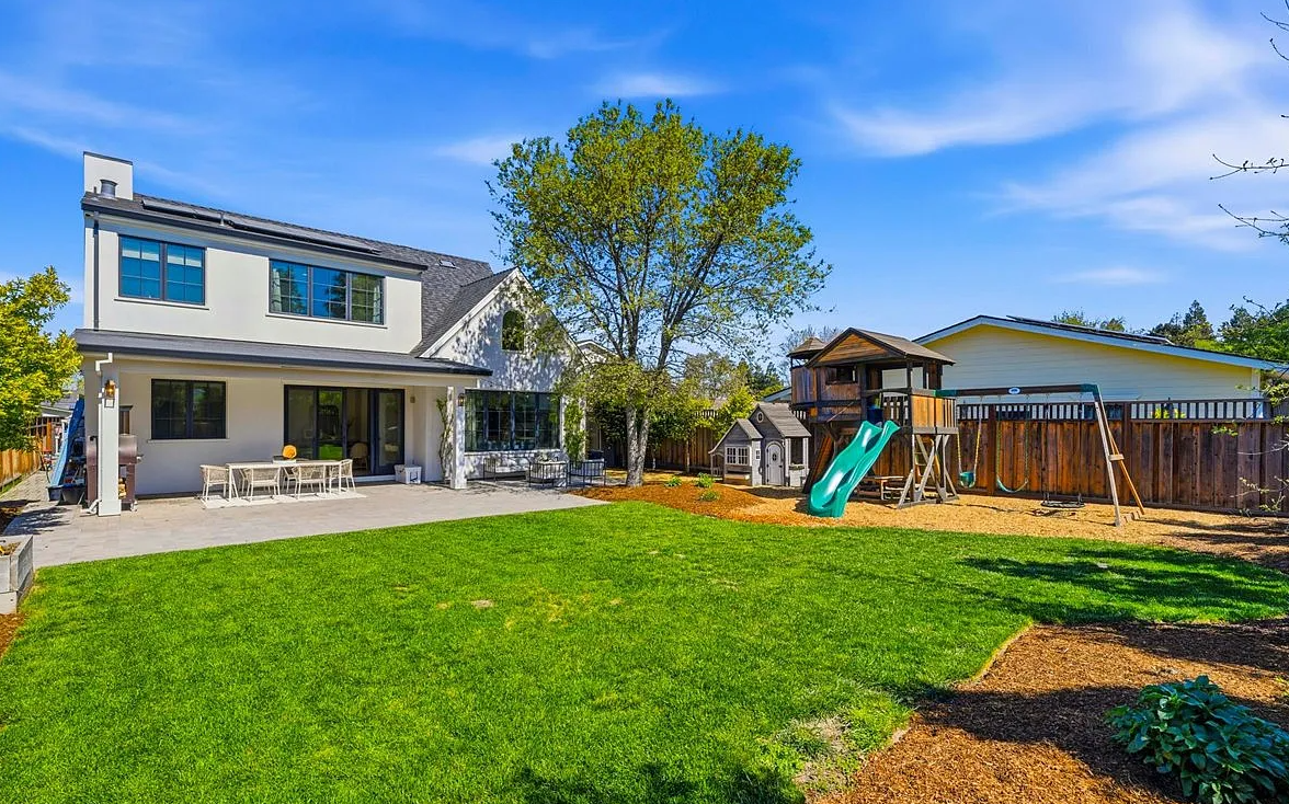 Modern two-story home with large backyard featuring green lawn, covered patio and wooden play structure.