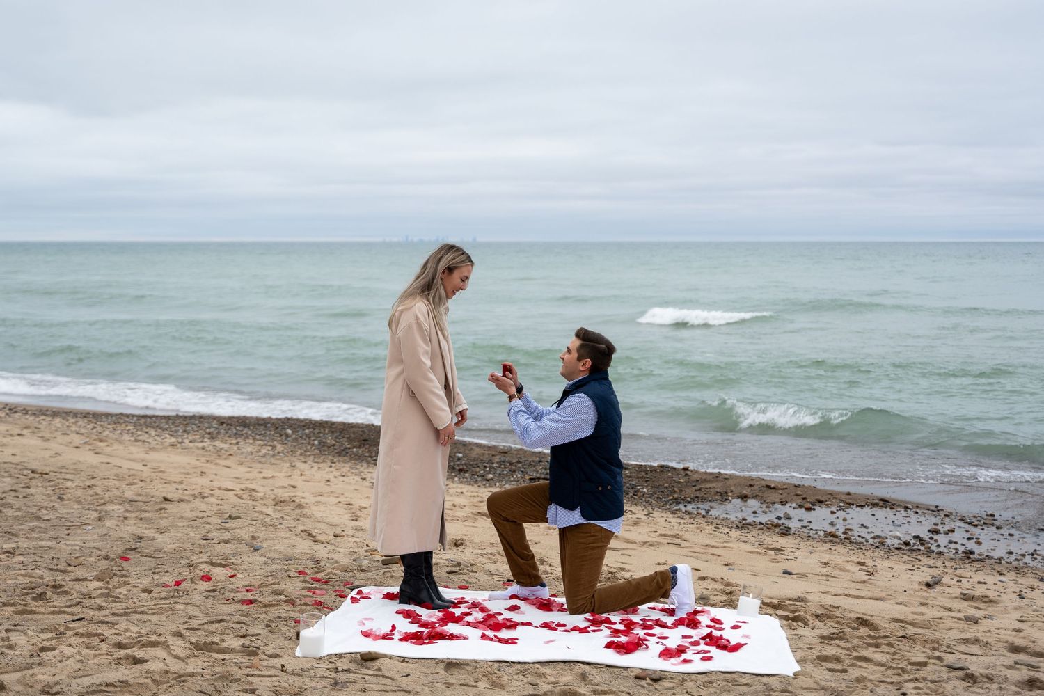 A romantic beach proposal scene with rose petals scattered on a white blanket by the ocean.