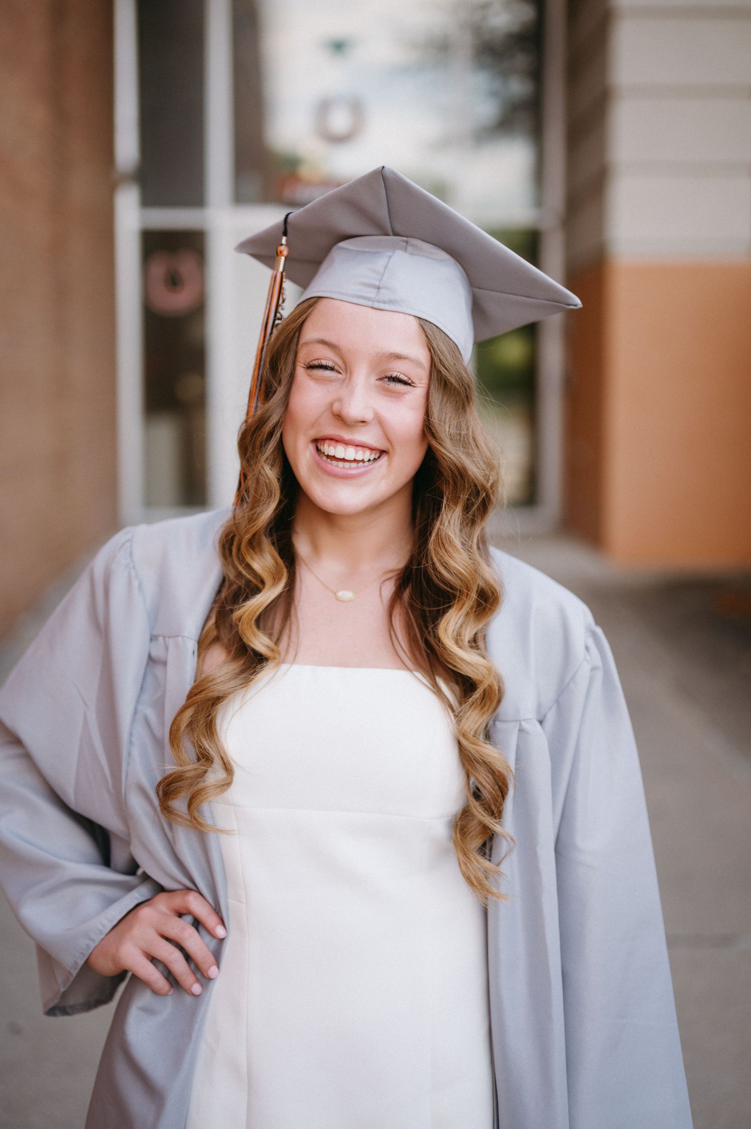 Graduate wearing silver cap and gown poses against neutral building background with joyful expressions.