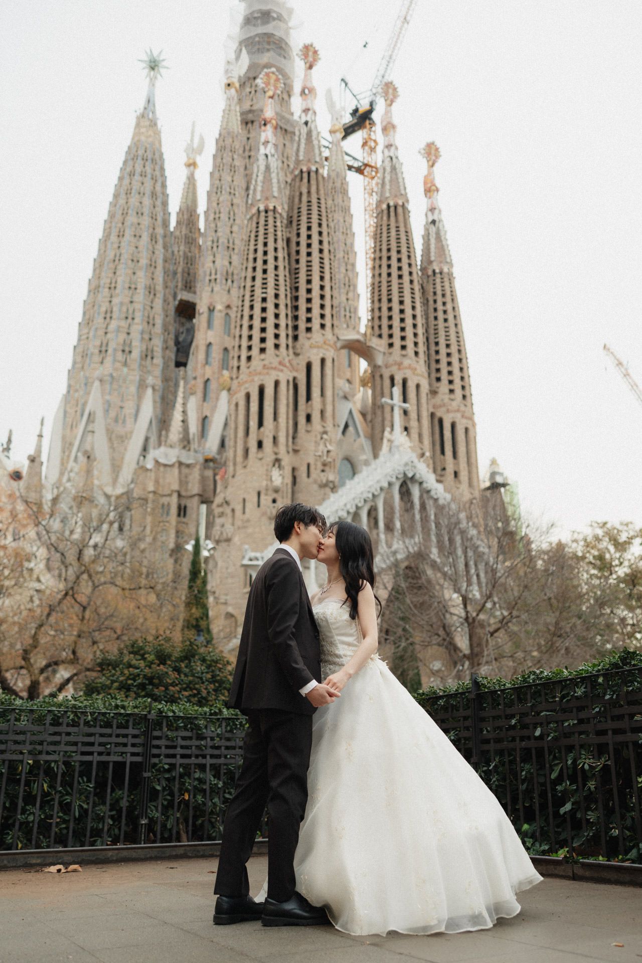 A couple in wedding attire share a romantic moment in front of the iconic Sagrada Familia cathedral in Barcelona, Spain.