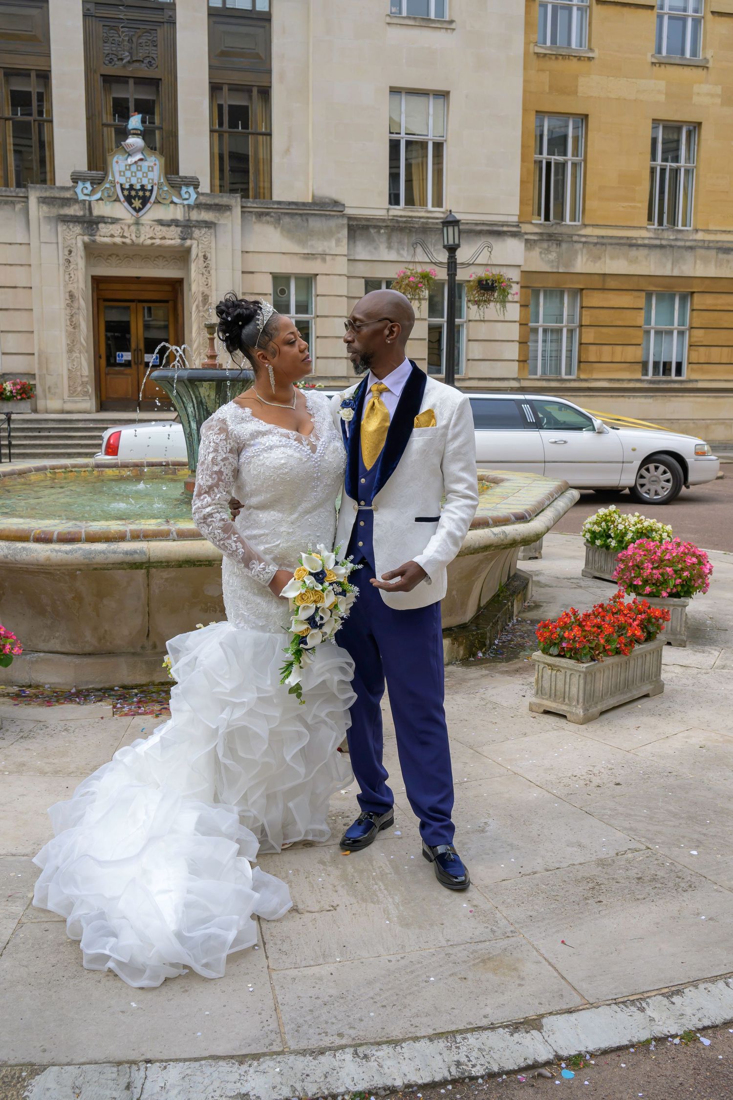 Elegant bride and groom posing by a stone fountain outside a grand building on their wedding day.