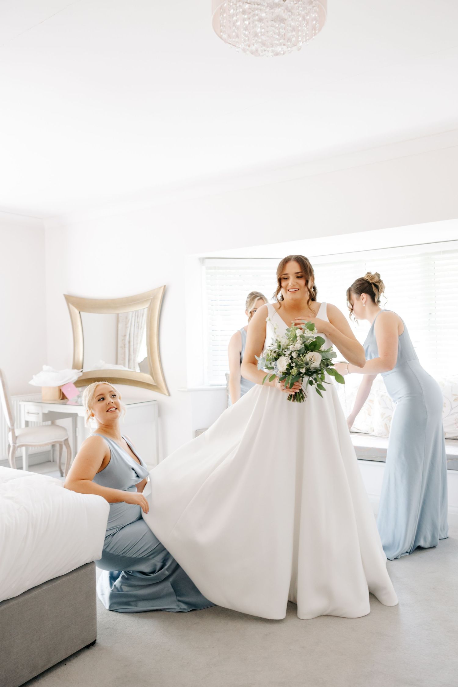 Bridesmaids in dusty blue dresses help adjust the bride's white wedding gown in a bright room at Wickham Estate wedding venue, Hampshire