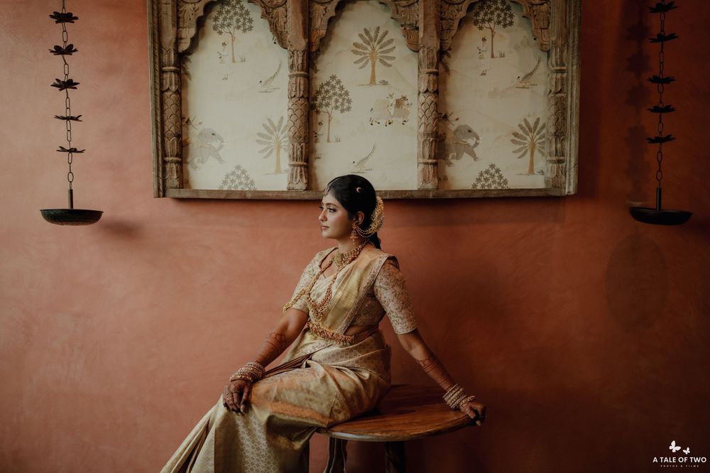Traditional South Asian bride in gold saree sitting on wooden bench against terracotta wall with decorative panels.