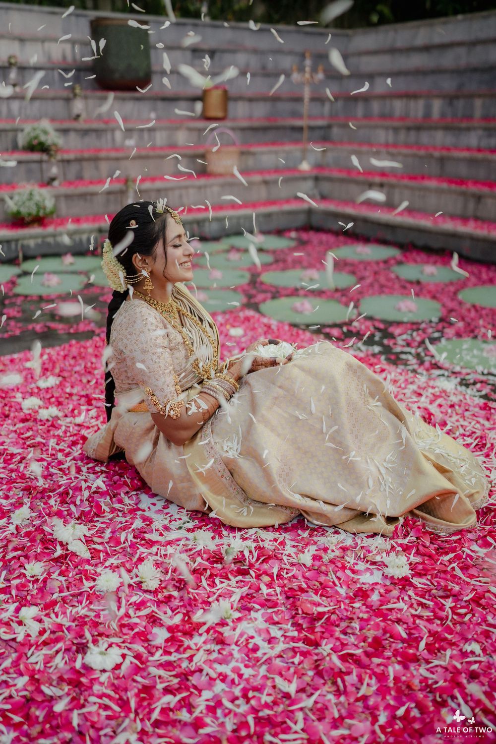 Traditional bride in gold and cream attire sits among scattered pink flower petals during a celebratory moment.