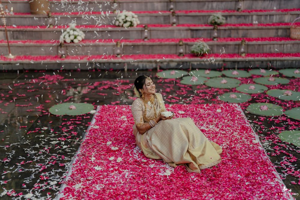 Artistic wedding photo on pink petal-covered mat with stone steps and scattered flowers in background.