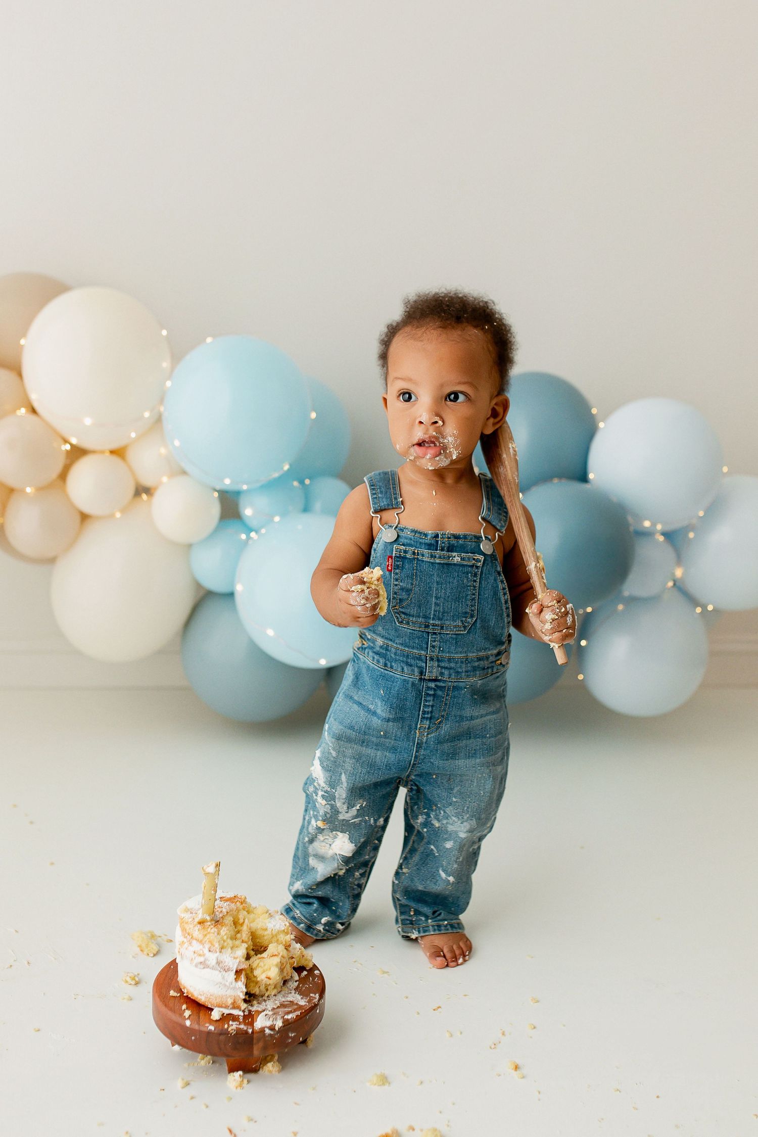 One year old boy wearing denim overalls with blue balloon arch with cake smash at Flowertown Photography Studio in Summerville, SC