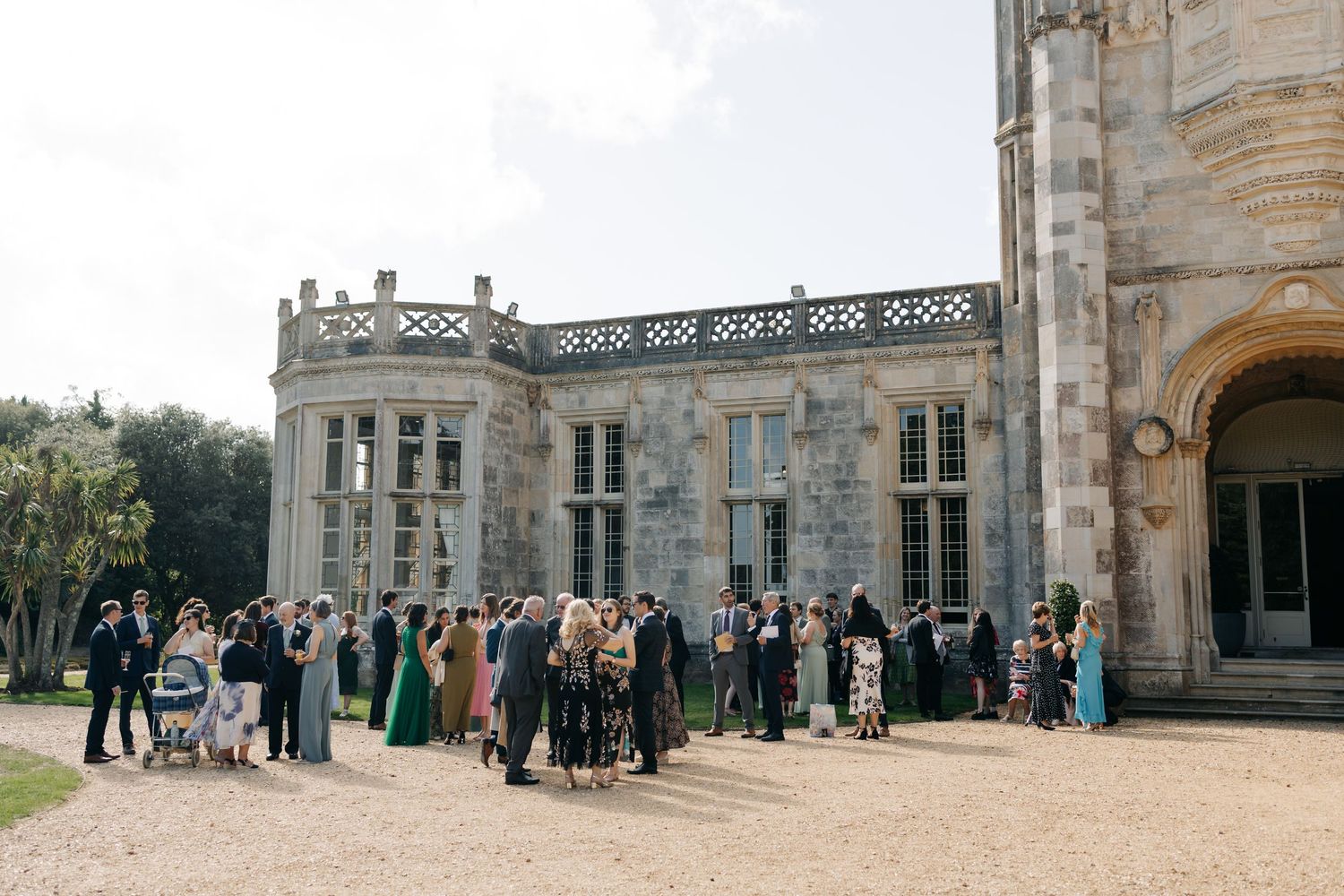 A panoramic view of wedding guests gathered outside a grand stone mansion during an elegant outdoor celebration.