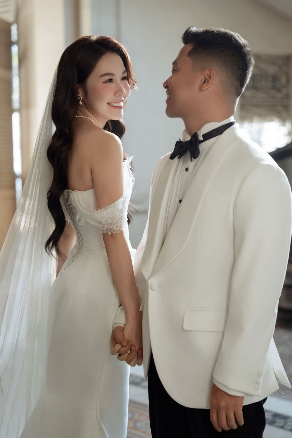 Bride and groom in white tuxedo hold hands smiling at each other in soft light.