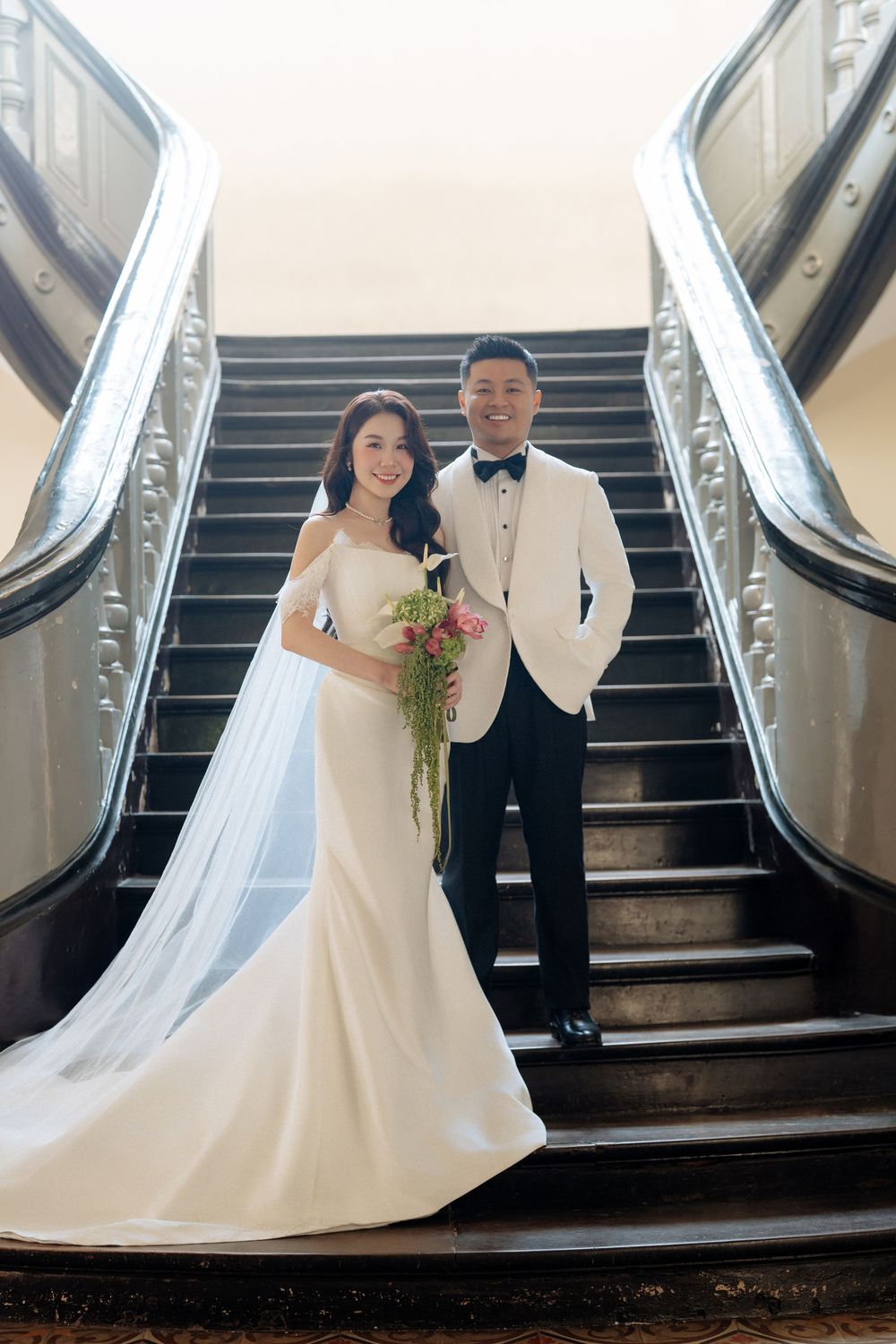 Bride in mermaid gown and groom in white tuxedo pose at base of grand staircase.