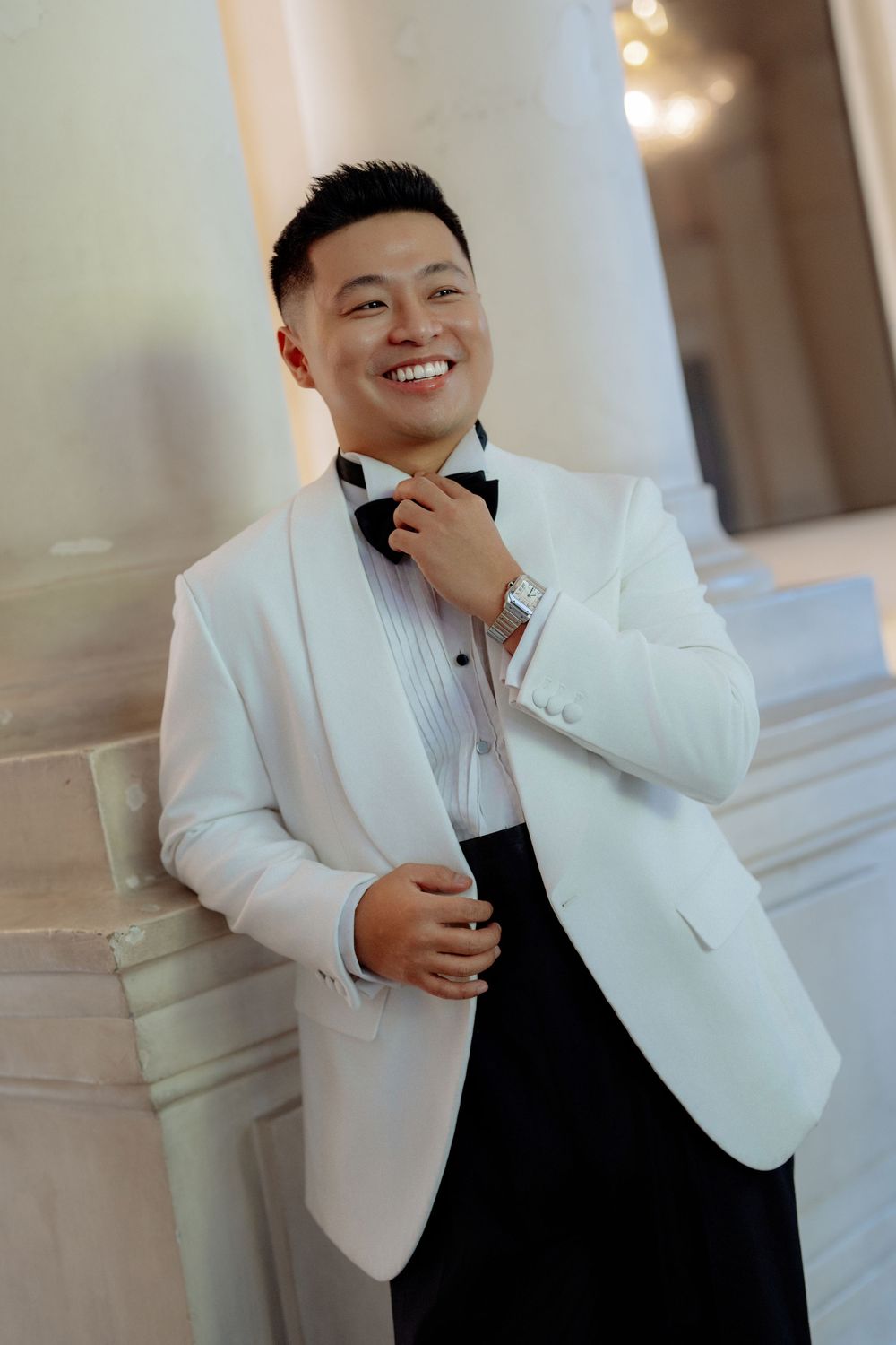 Smiling groom in white tuxedo jacket adjusts bow tie leaning against marble column.