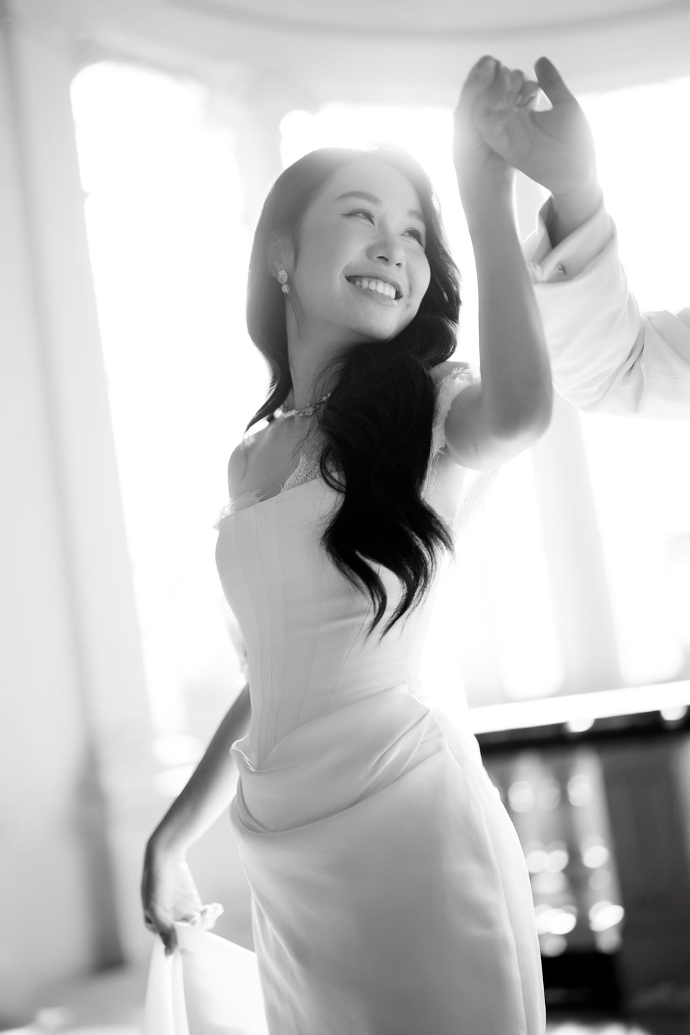 Black and white photo of joyful bride in white gown dancing in sunlit room.