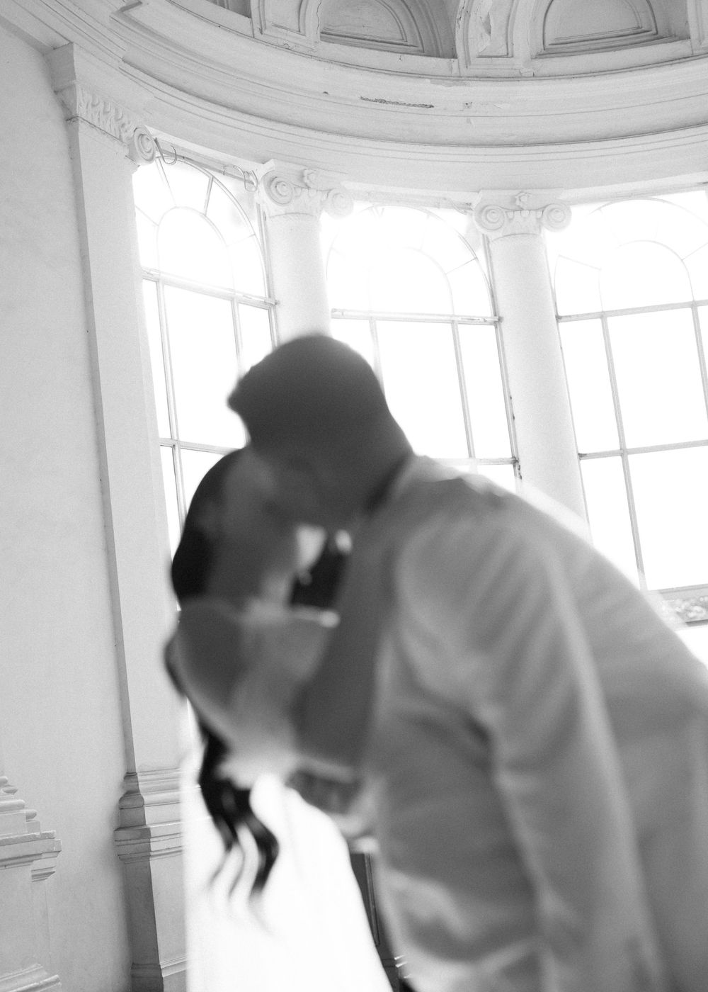 Black and white artistic blur of bride and groom kissing before large arched windows.