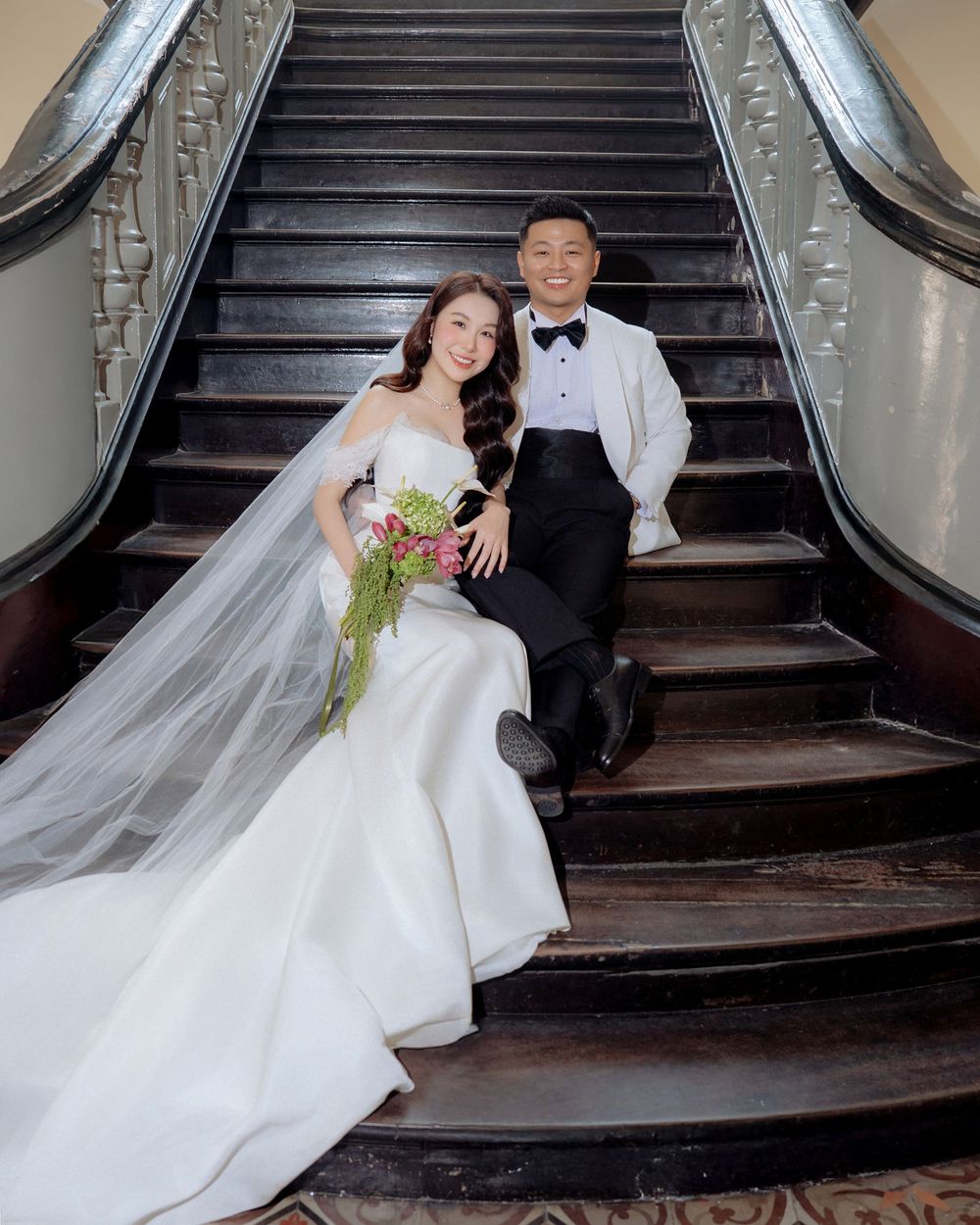 Bride and groom sit together on ornate dark wood staircase with cascading bridal bouquet.