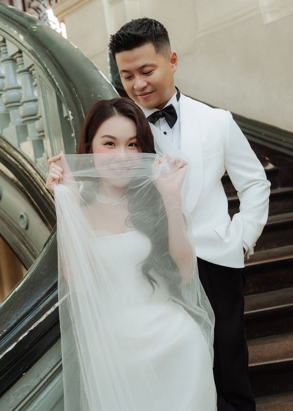 Bride holds veil over face while groom in white tuxedo smiles behind her on elegant staircase.