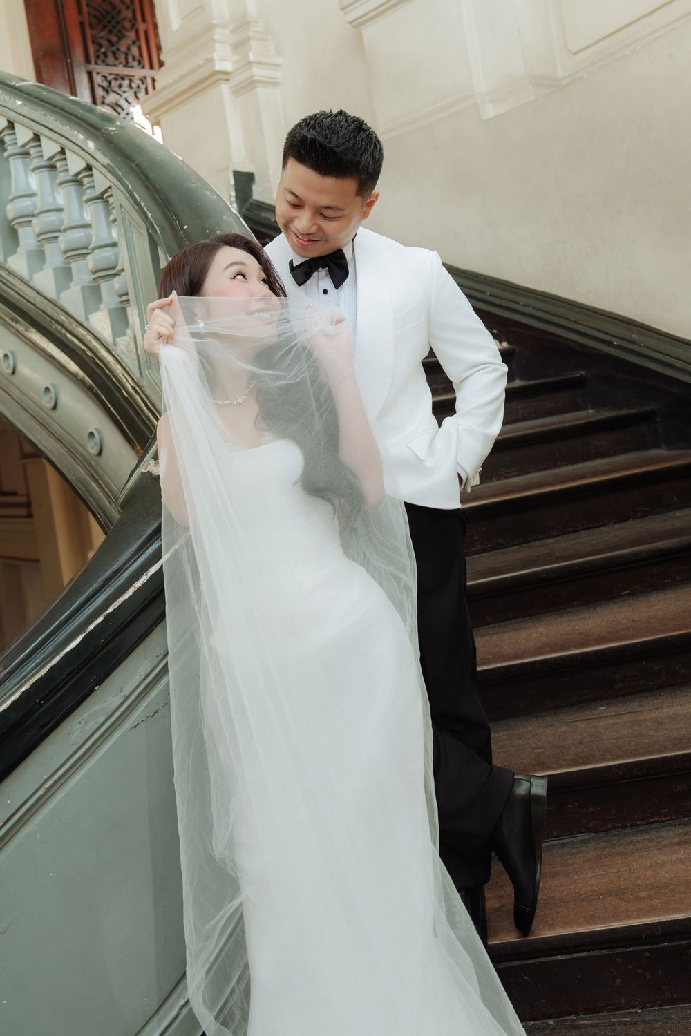 Wedding couple poses on grand staircase, bride in white gown with flowing veil, groom in white tuxedo.