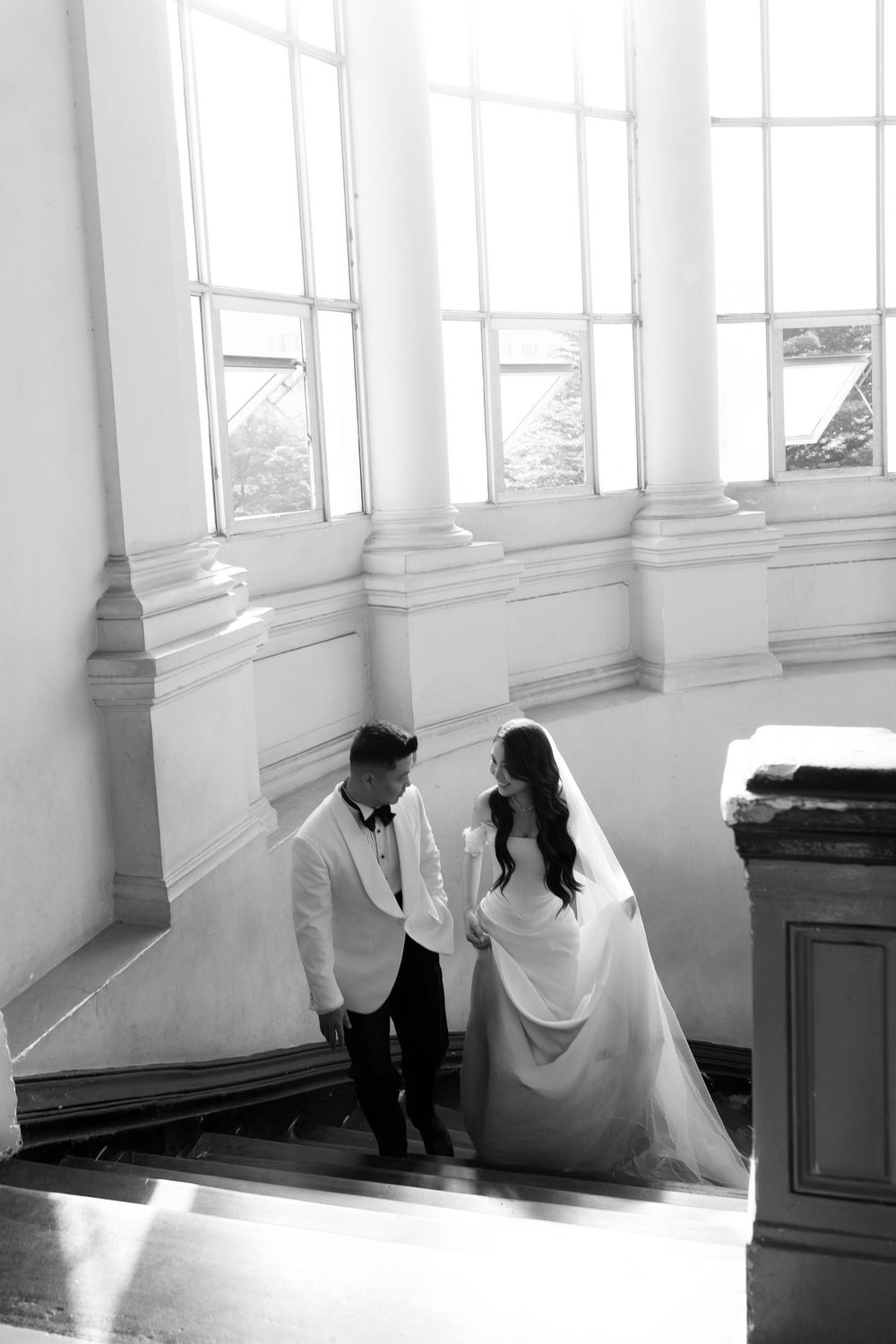 Black and white photo of bride and groom holding hands on sunlit staircase with tall windows.