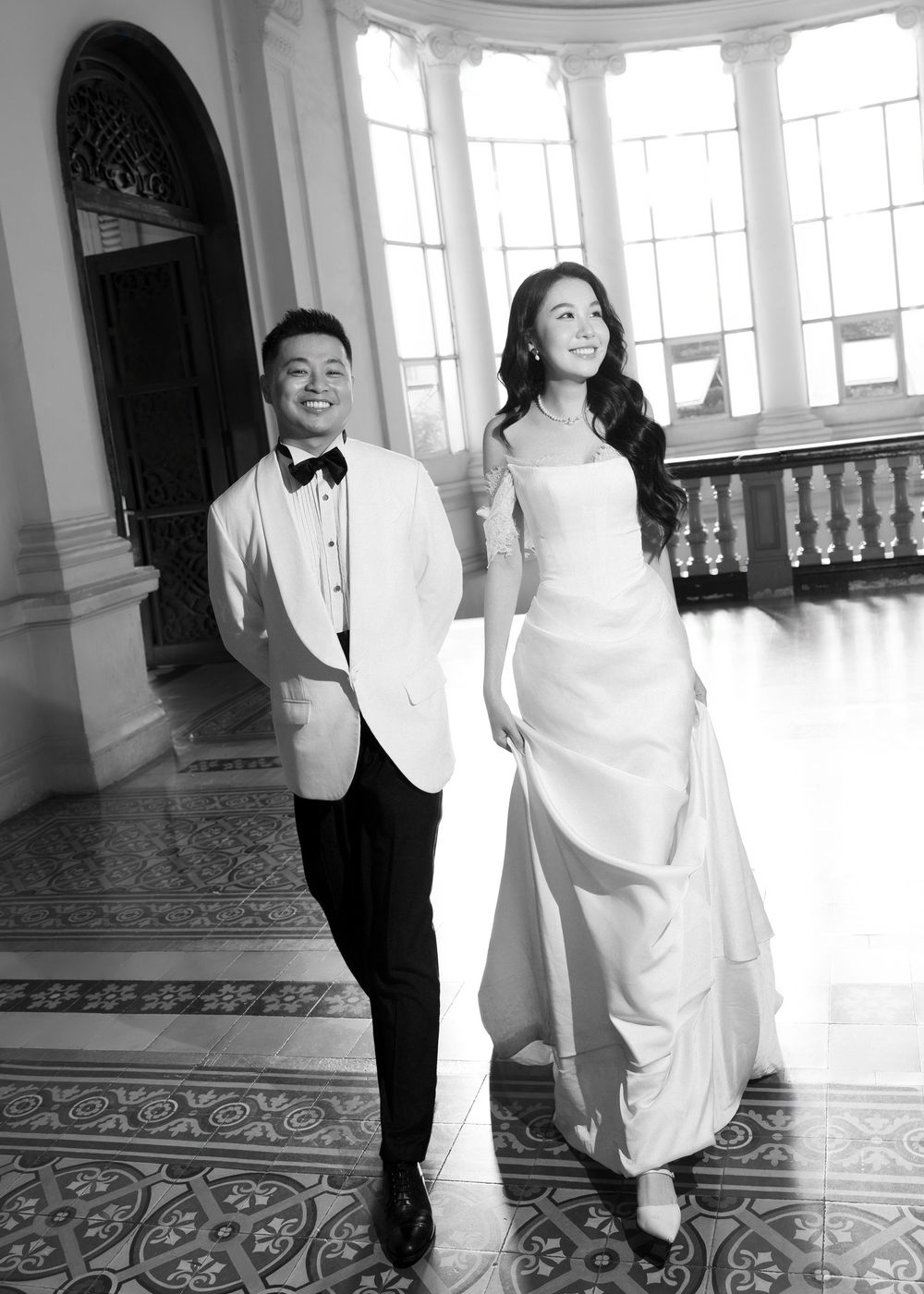 Elegant black and white photo of Asian couple in wedding attire walking in grand architectural space.