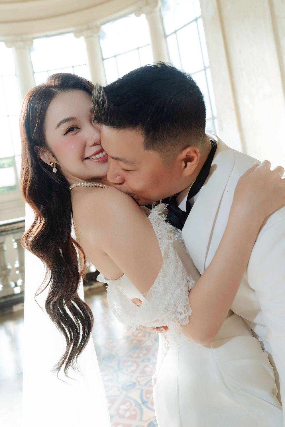 Groom in white tuxedo kisses smiling bride on cheek during romantic wedding photo session.