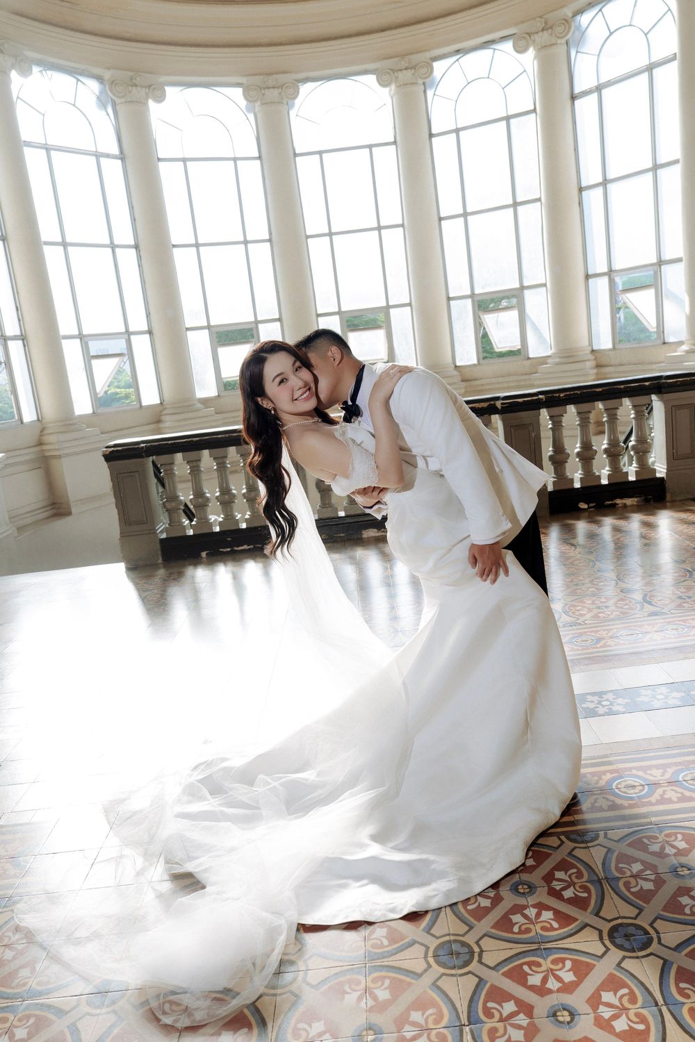 Bride and groom dance together in white gown and tuxedo in sunlit rotunda with ornate windows.