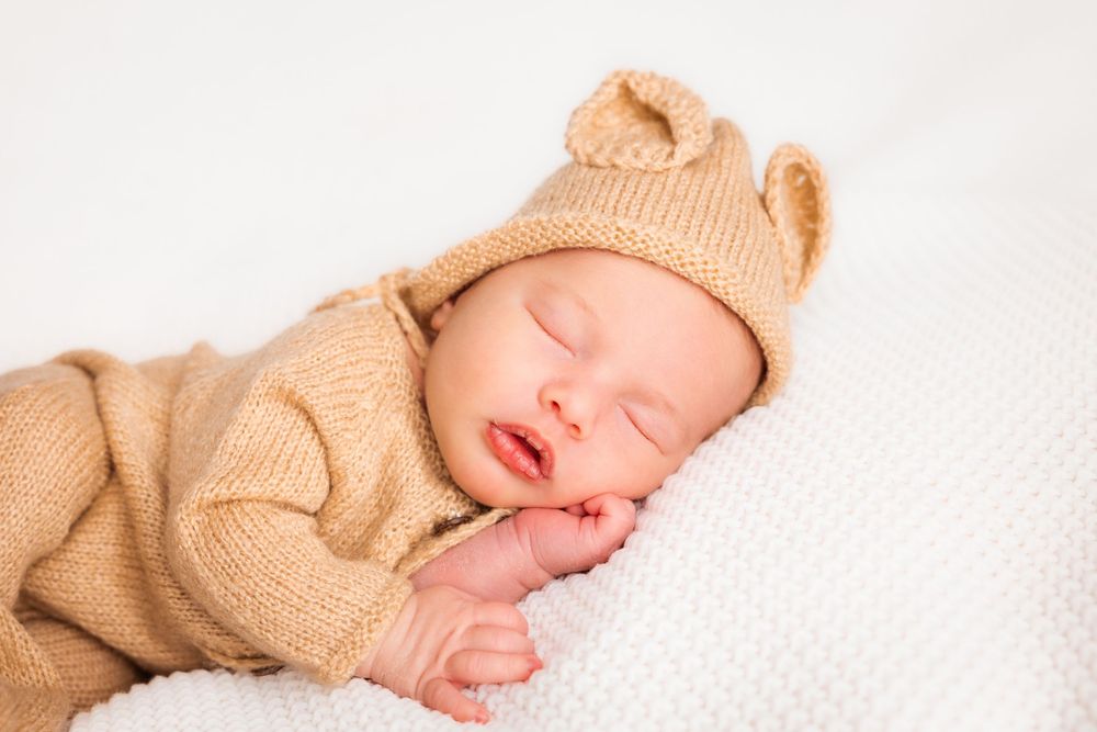 Sleeping baby wearing a cozy beige knit sweater rests peacefully on white blankets.