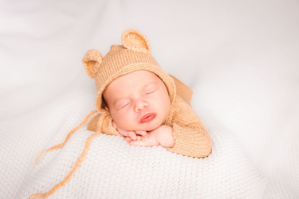 Sleeping newborn baby wearing peach-colored knit hat rests peacefully on soft white blanket.