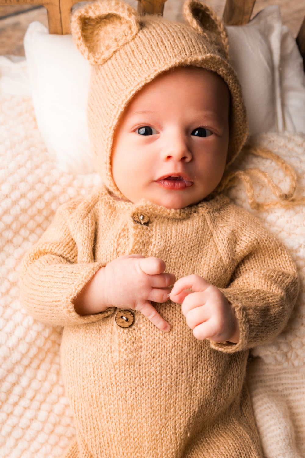 A small baby wearing a cozy beige knit sweater and matching hat lies on a textured white blanket.