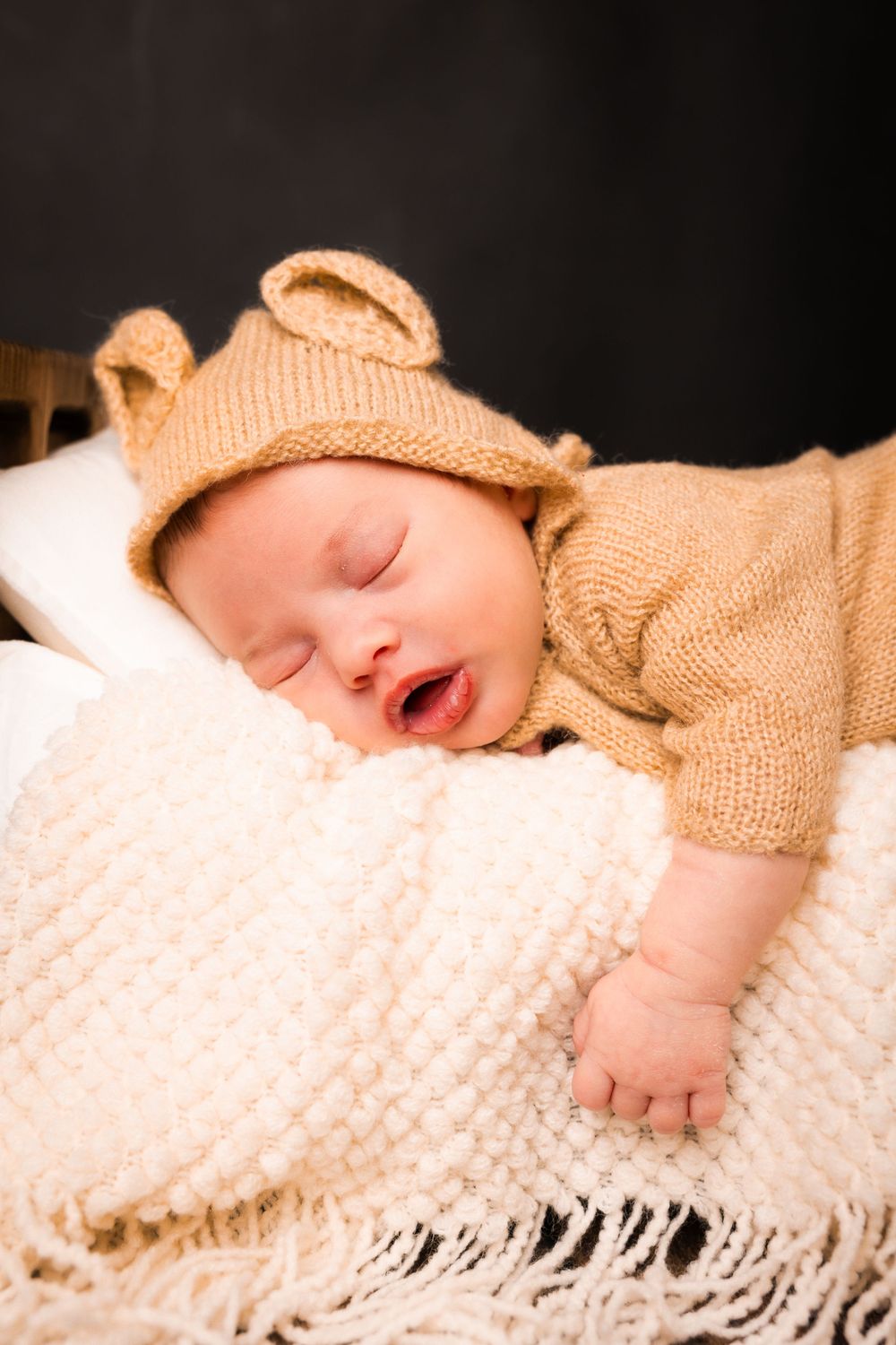 A sleeping baby wrapped in a cozy beige knit blanket lies peacefully on white textured bedding.