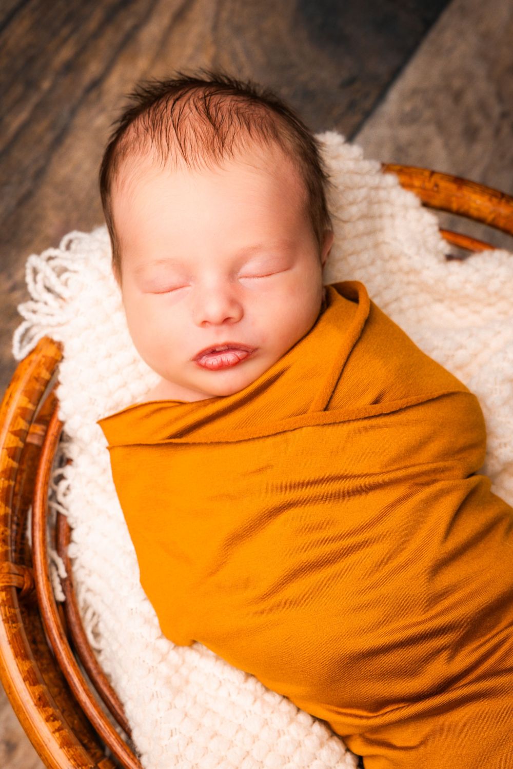 A sleeping newborn baby wrapped in a cozy orange swaddle blanket rests peacefully on white textured fabric.