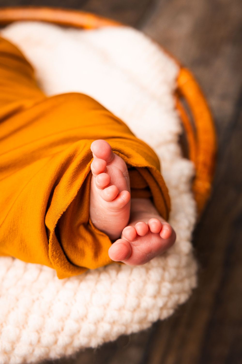 A tiny baby foot peeks out from under an orange blanket on a white knitted surface.