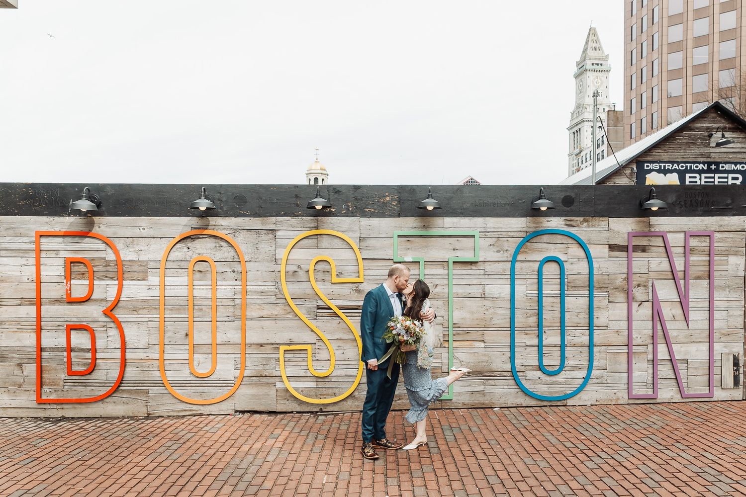 Couple sharing a romantic kiss in front of the colorful Boston sign mural on a brick wall in Boston, MA.