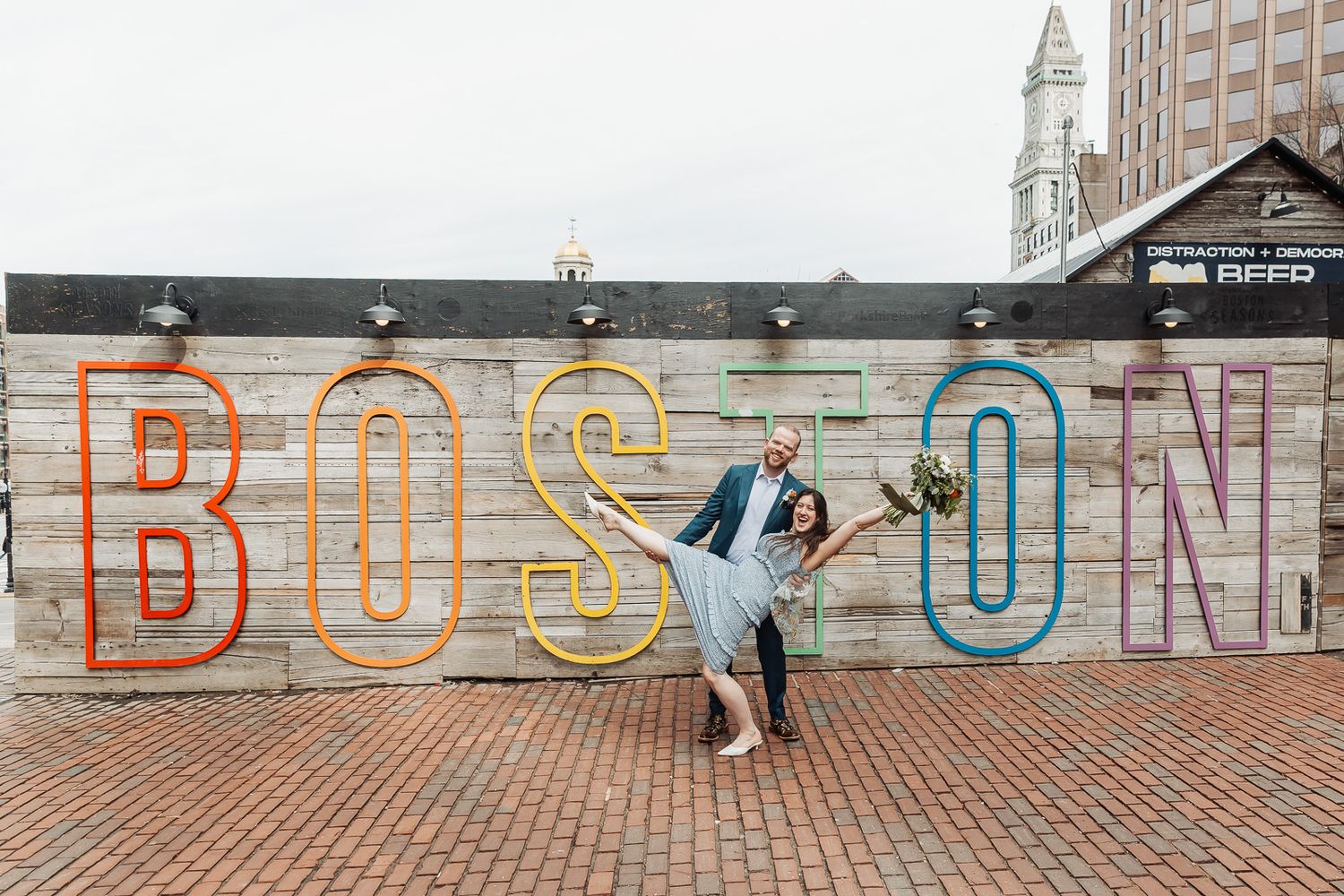 Couple dancing joyfully in front of colorful Boston sign mural on brick wall, wedding attire, bouquet.