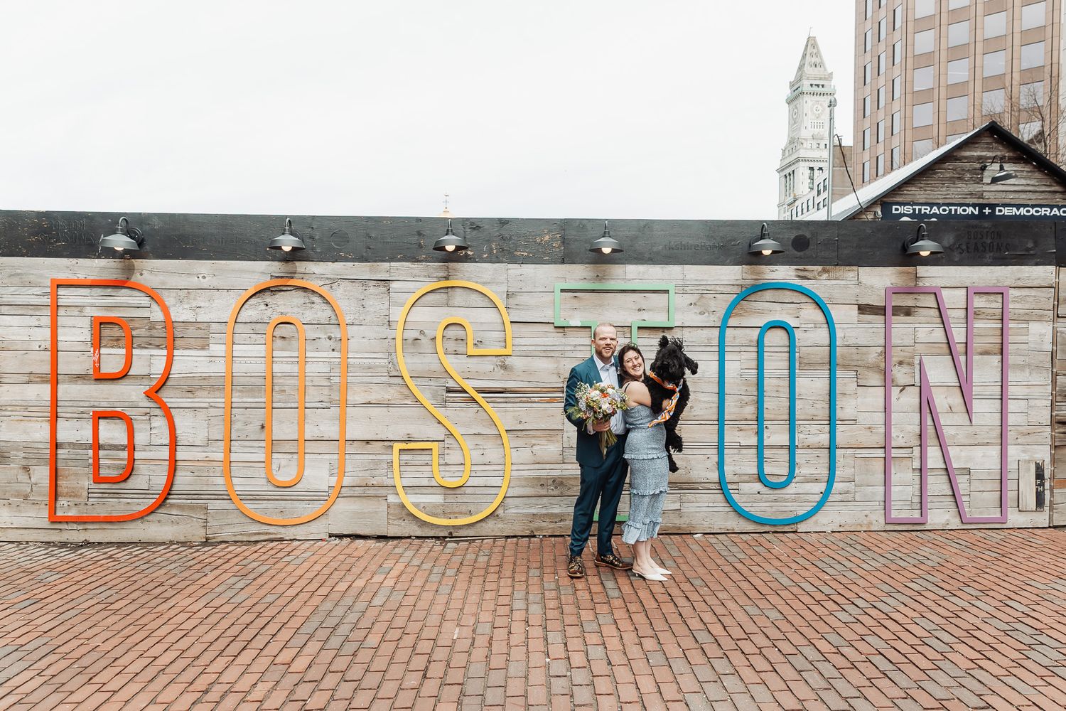 Family posing in front of colorful BOSTON sign mural on brick plaza with city skyline in background.