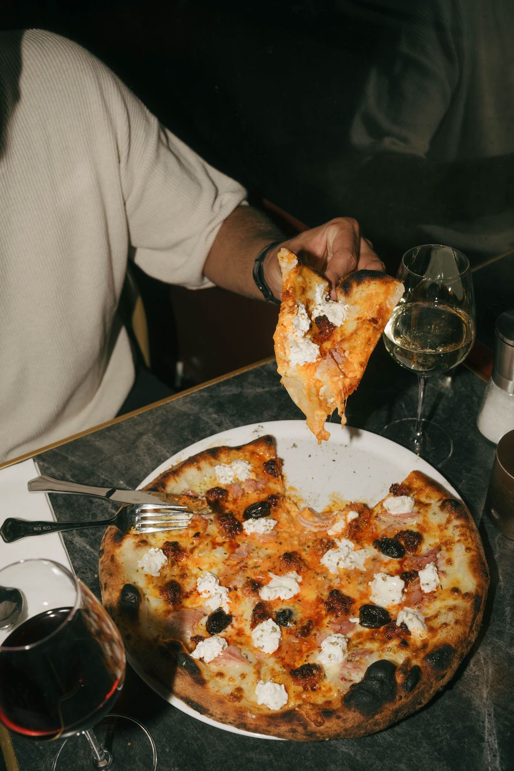 Personne soulevant une tranche de pizza au fromage d'une assiette blanche, avec des verres à vin sur une table de restaurant sombre.
