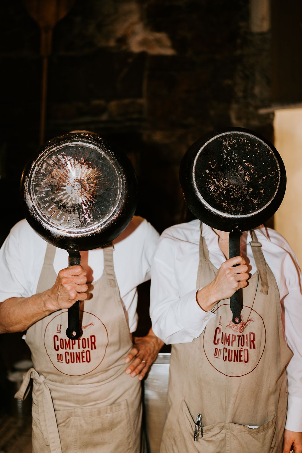 Deux chefs dans les tabliers du Comptoir de Cuneo cachent avec humour leurs visages derrière des poêles à frire bien utilisées.