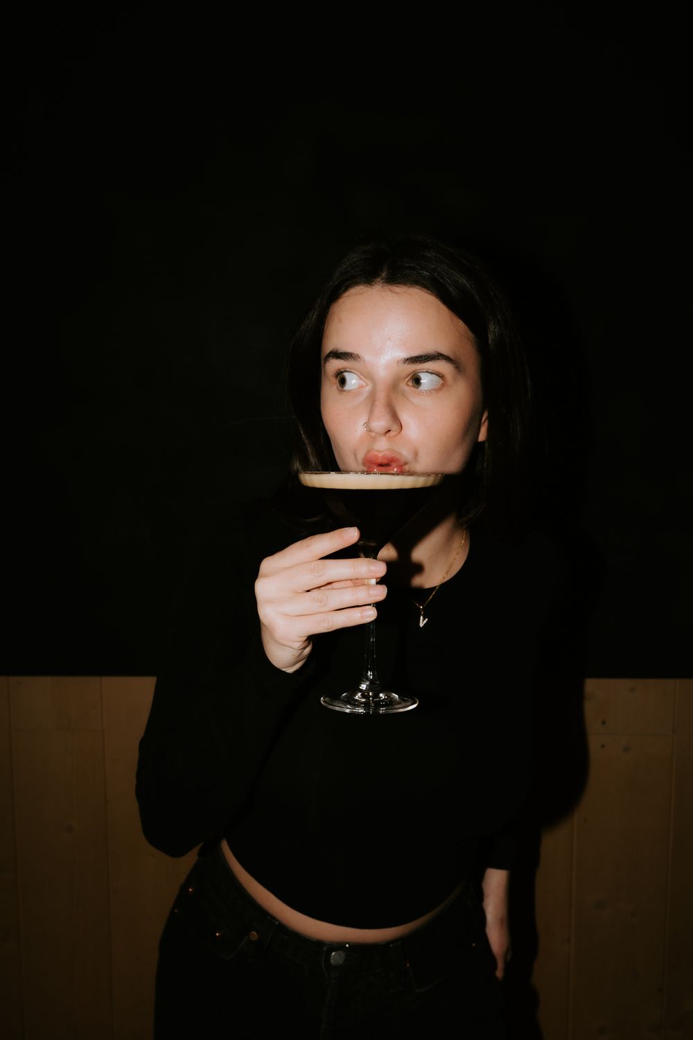 Jeune femme en haut court noir sirotant un cocktail sombre dans un verre à tige sur fond sombre.