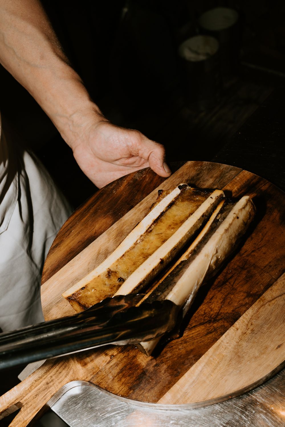 Chef présentant de la moelle osseuse rôtie sur une planche à découper en bois rustique, garnie d'herbes, dans un cadre de restaurant sombre.