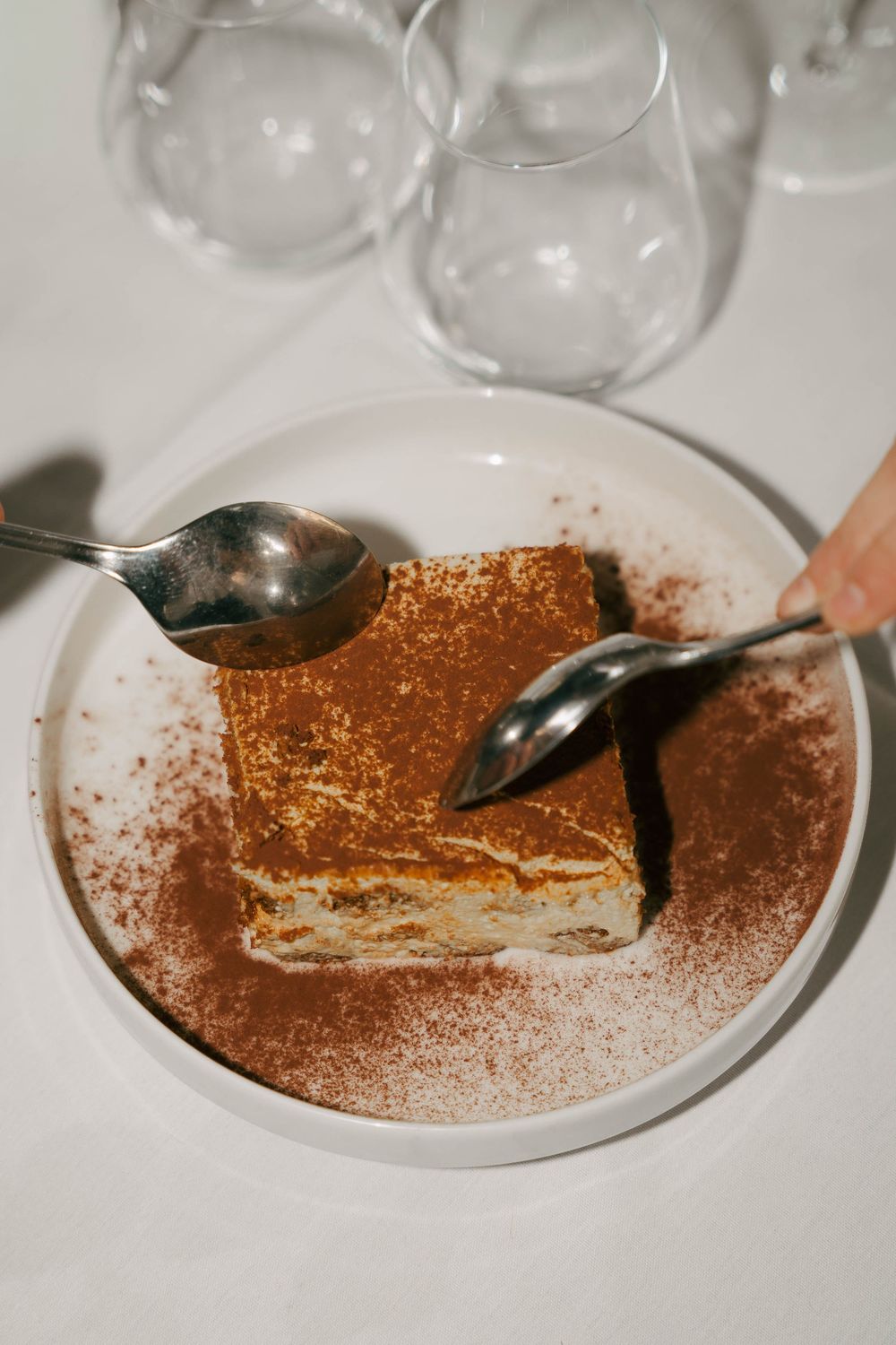 Une tranche de tiramisu saupoudrée de poudre de cacao sur une assiette blanche, avec deux cuillères et des verres à vin en arrière-plan.